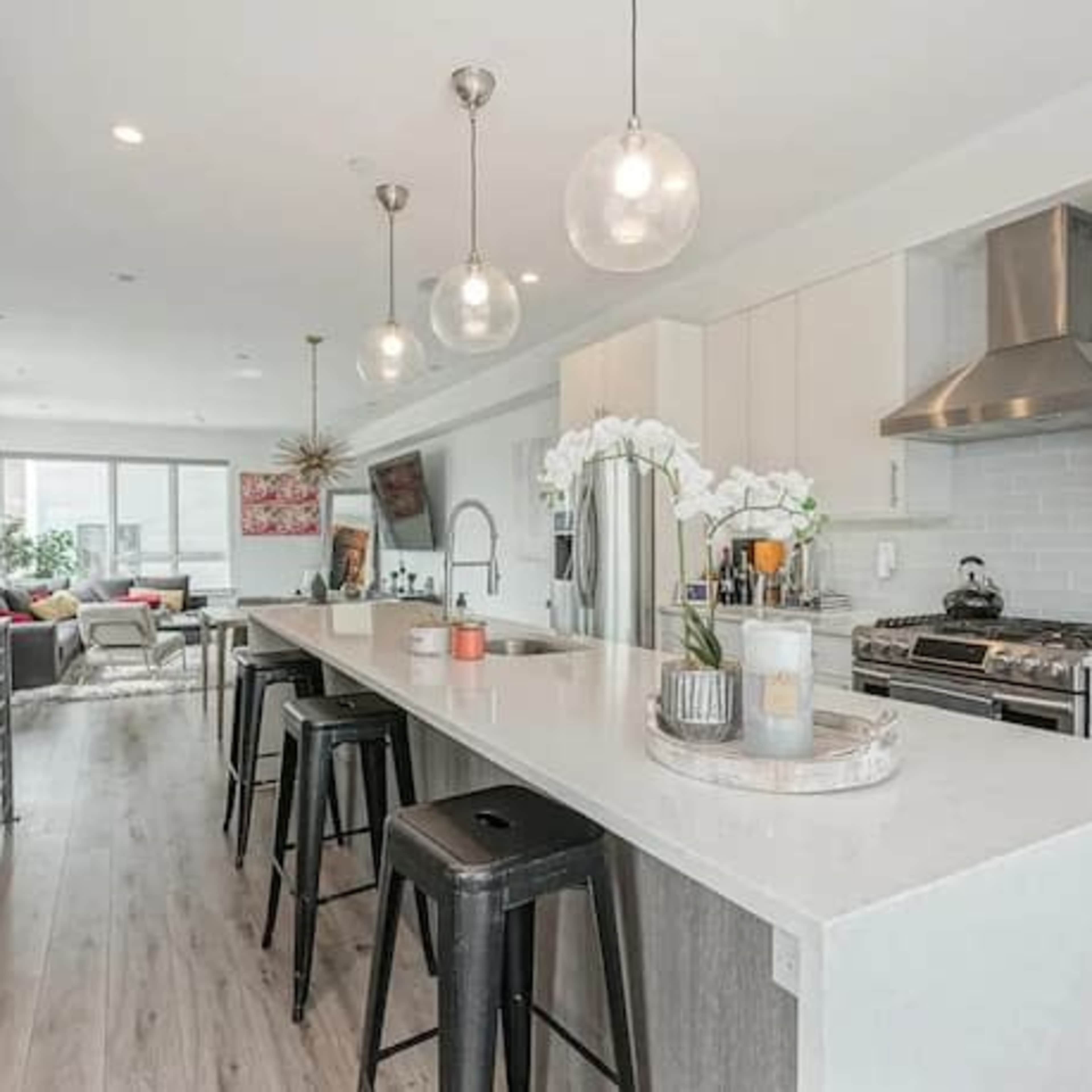 A modern kitchen and living area with a long white island, bar stools, stainless steel appliances, and large windows.
