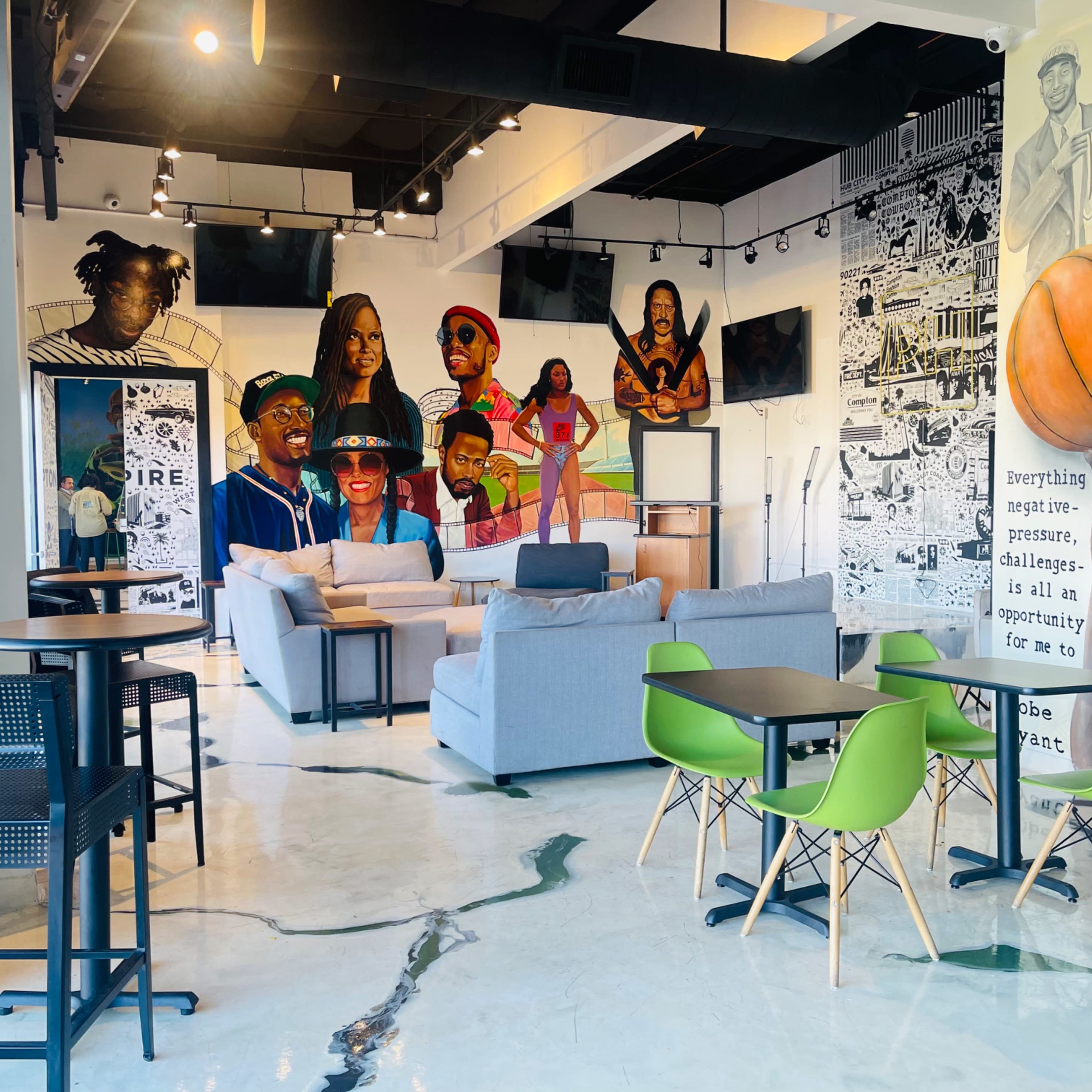 A modern indoor space featuring gray sofas, green chairs, and a large mural with diverse figures, including athletes, on the walls.