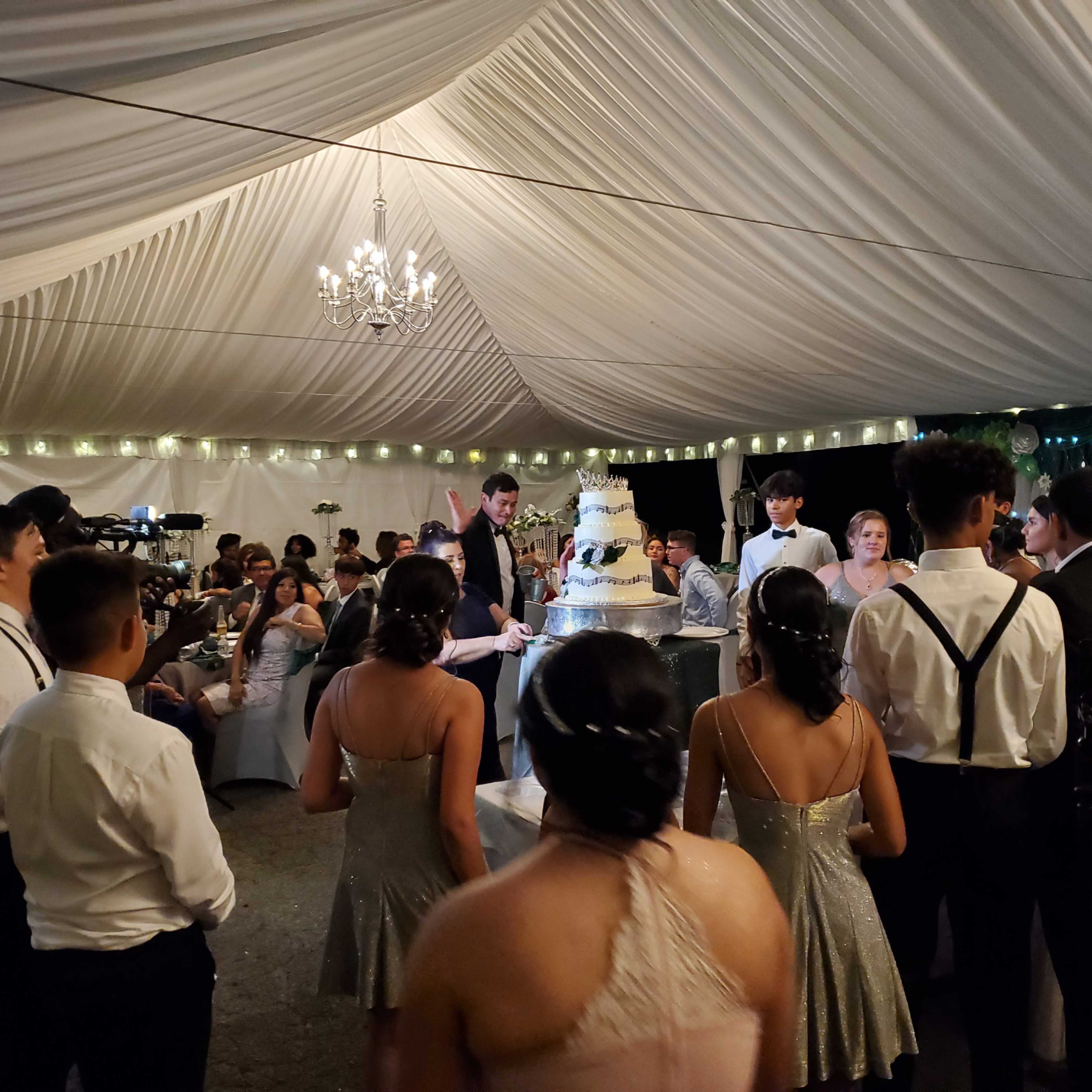 A group of people gathered under a decorative tent watches as a couple prepares to cut a large wedding cake.