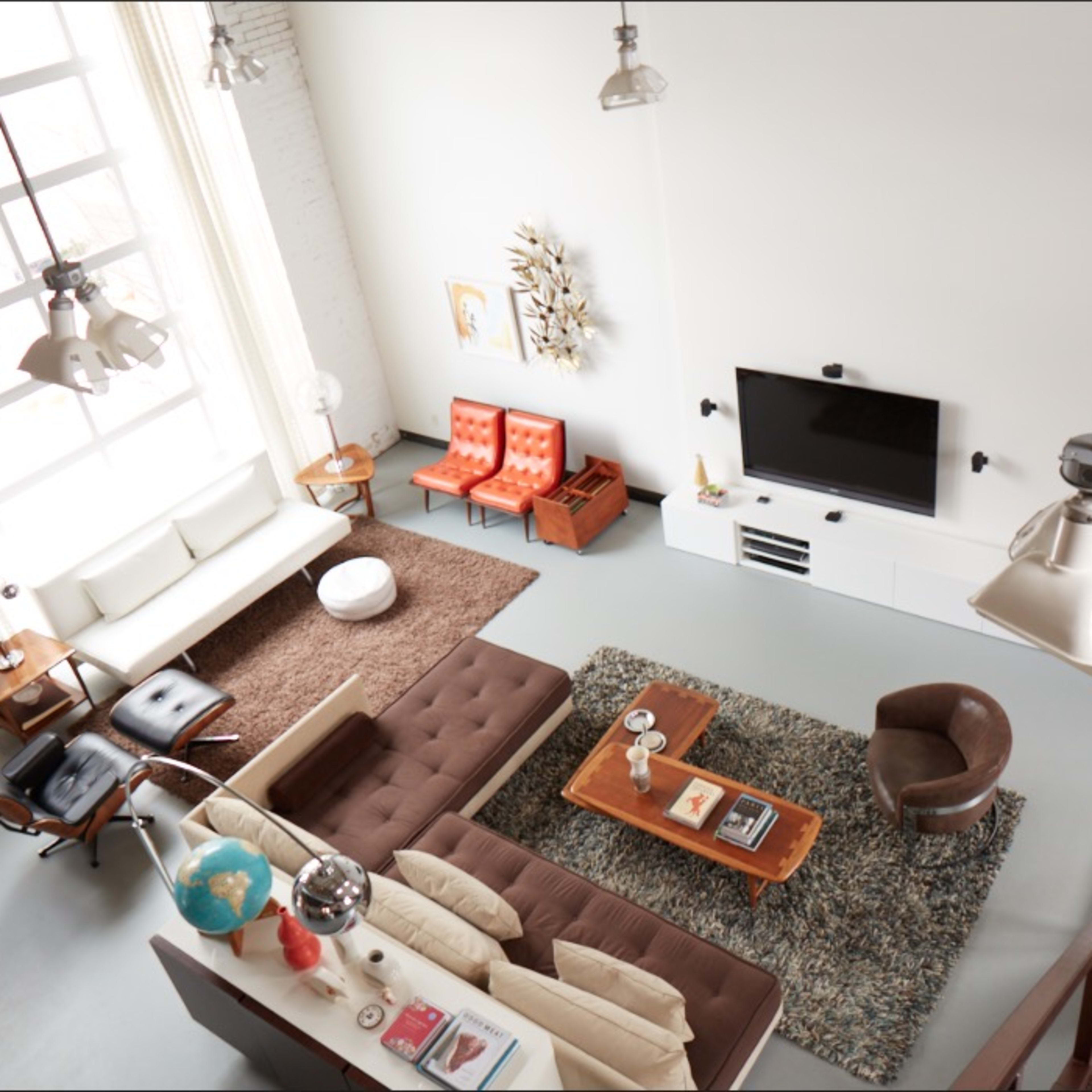 A modern living room with a large window, featuring two orange chairs, a brown sofa, a coffee table, and a television mounted on the wall.