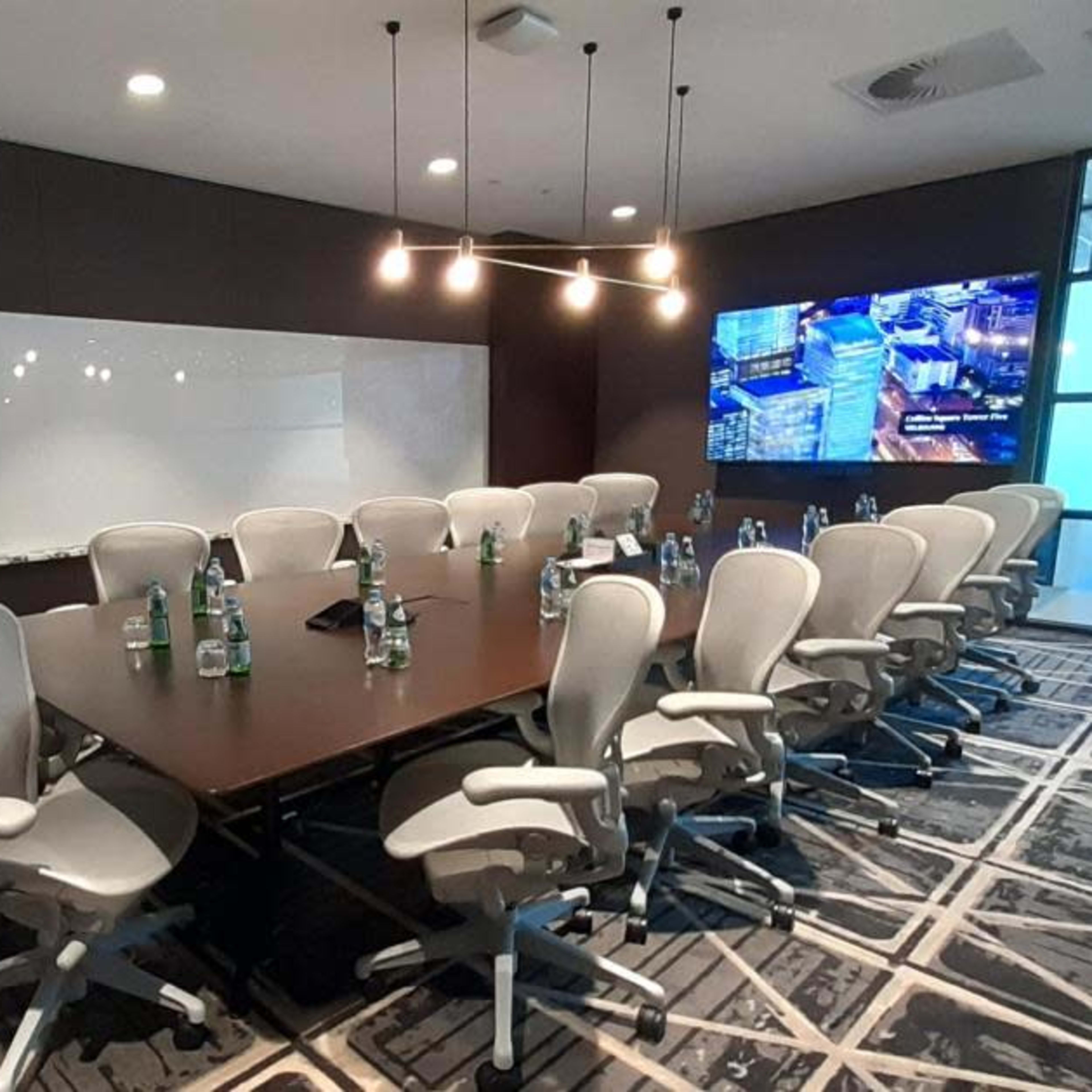 A large conference room features a long table surrounded by ergonomic chairs, a whiteboard, and a screen displaying an image.