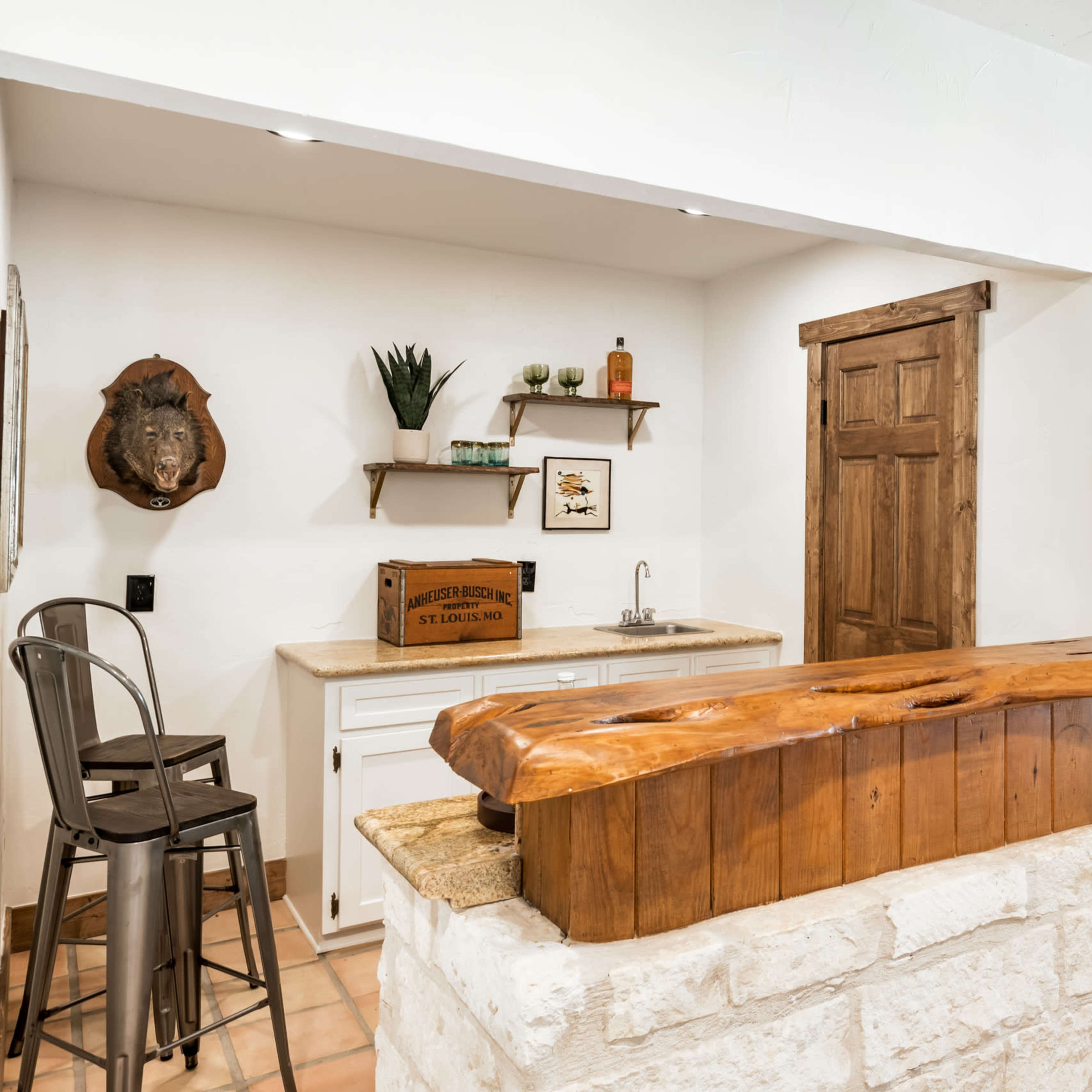 A rustic kitchen with a wooden bar counter, a sink, and a wooden door, complemented by stone and tiled flooring.
