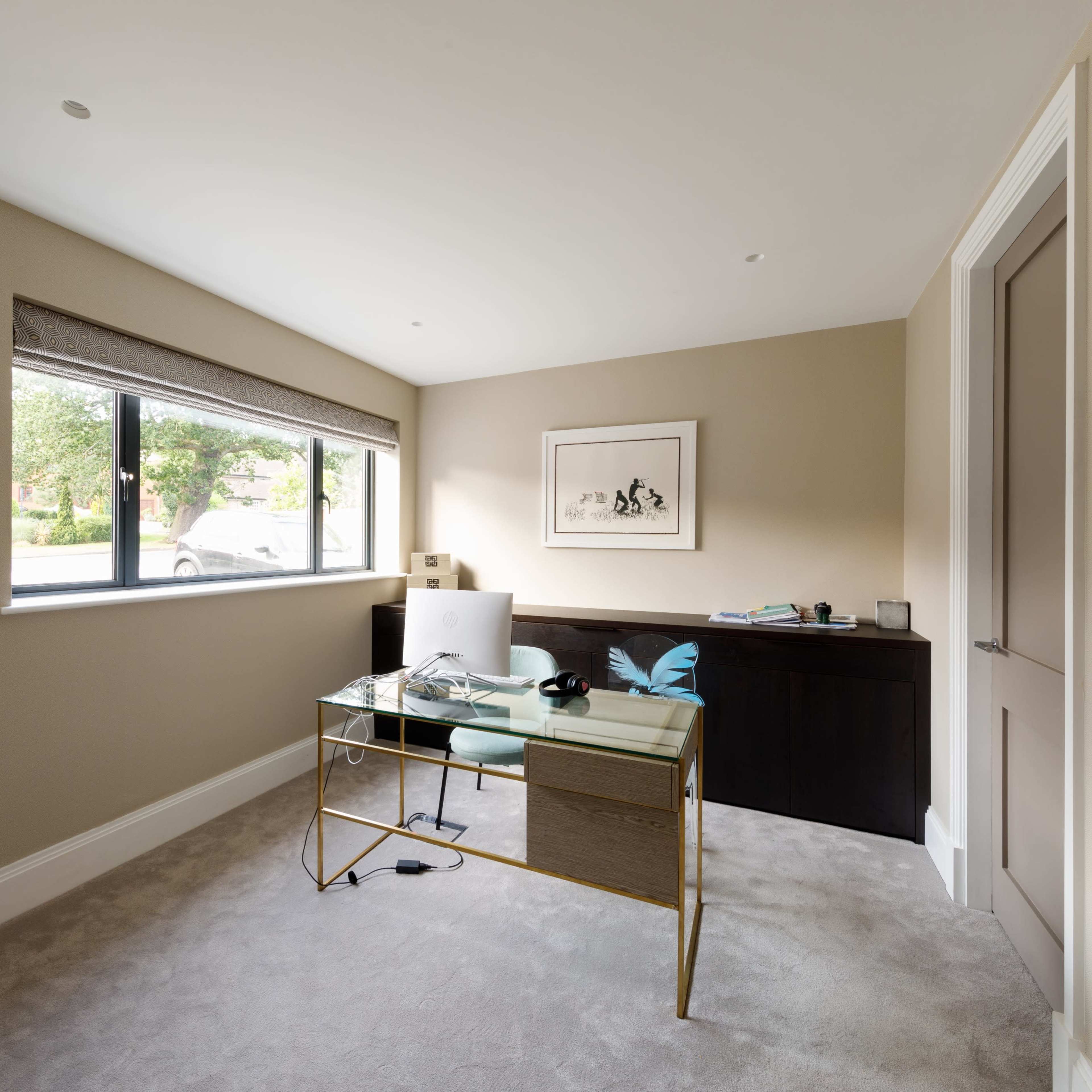 A minimalist home office with a glass-top desk, a chair, and a dark wooden cabinet, illuminated by natural light from a nearby window.