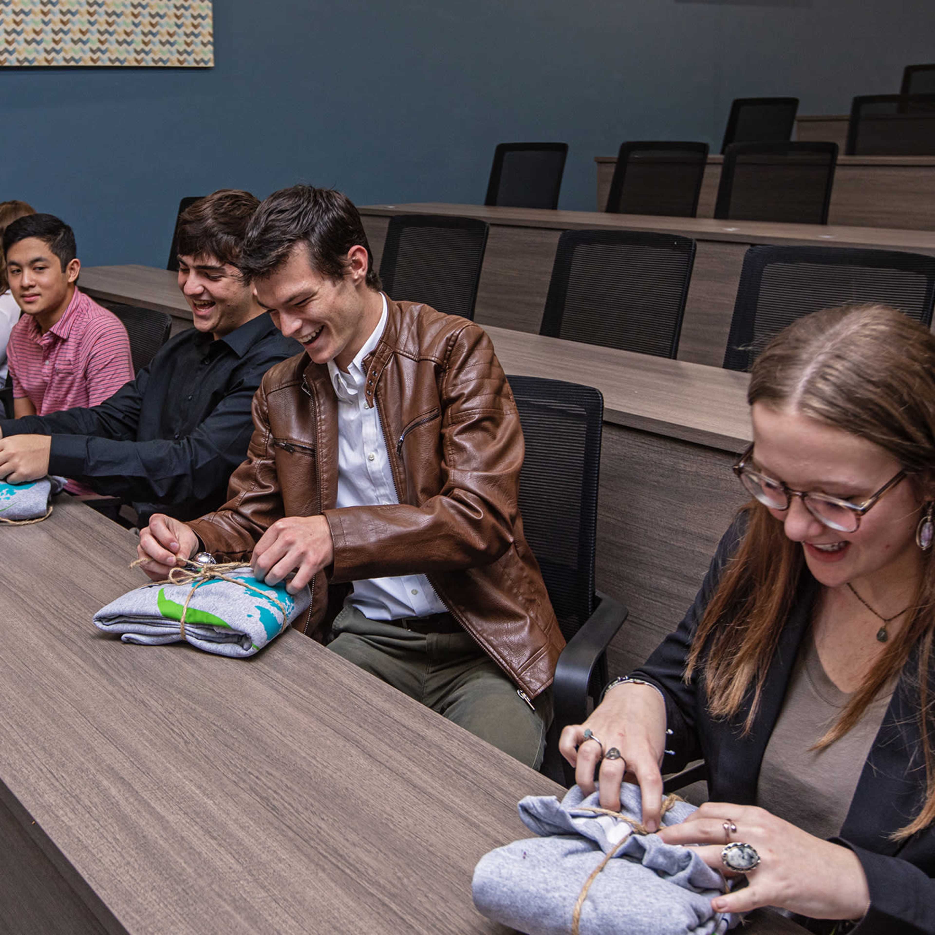 A group of six people sits at a long table, wrapping clothing items in a conference room.