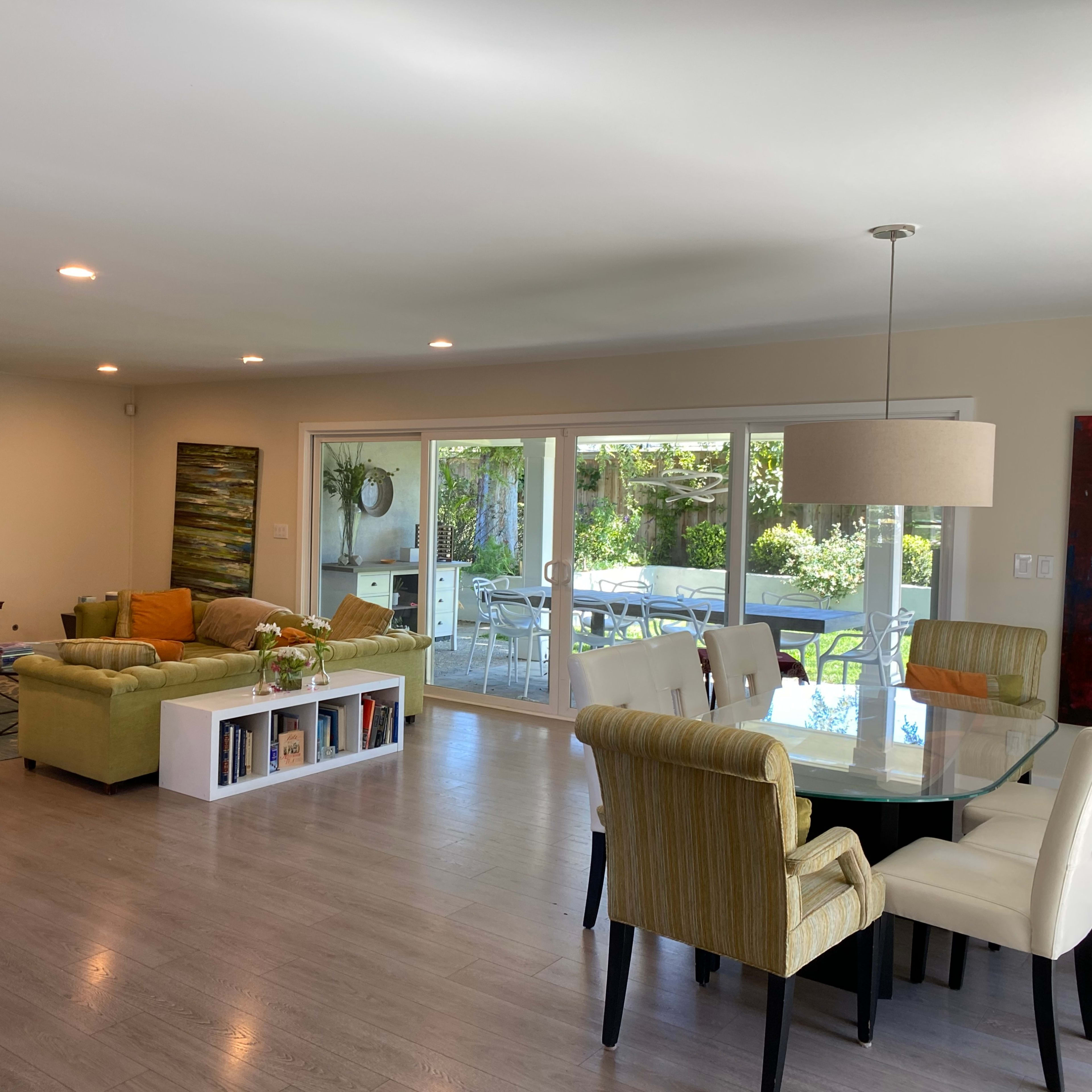 A spacious living area with a green sofa, a dining table, a fireplace, and large windows overlooking an outdoor patio.