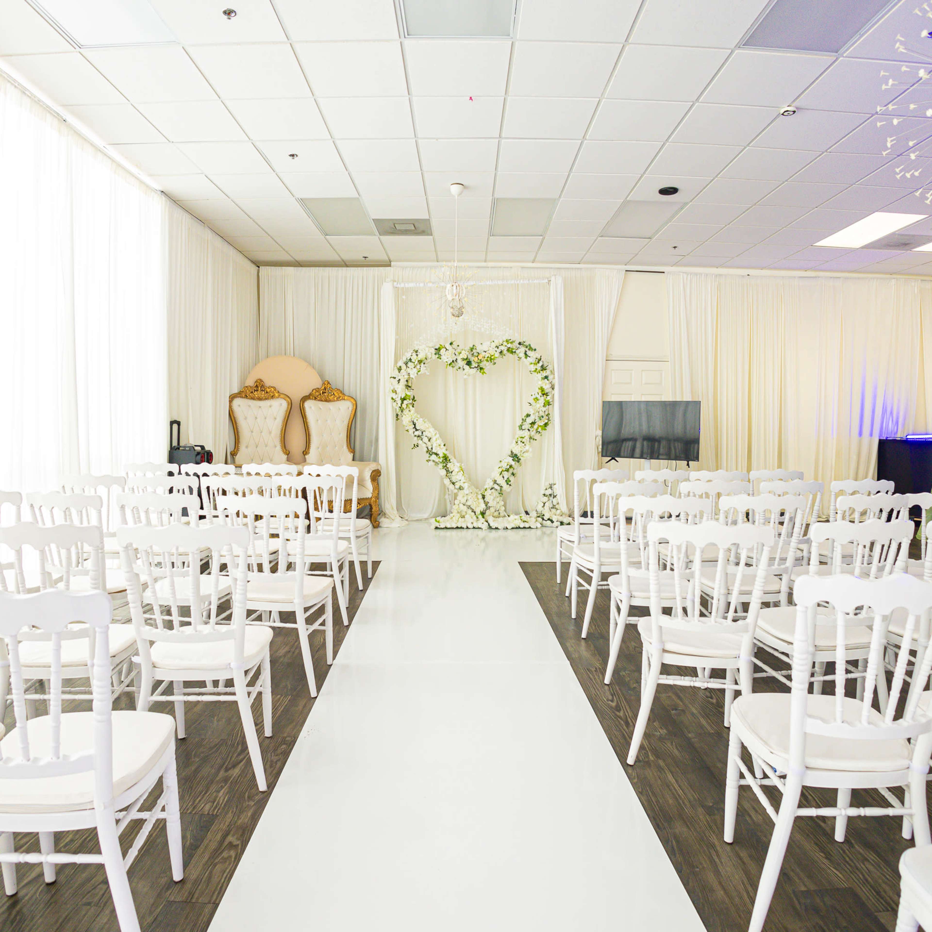 A wedding ceremony setup with rows of white chairs facing a floral heart arch in a bright room with sheer curtains.