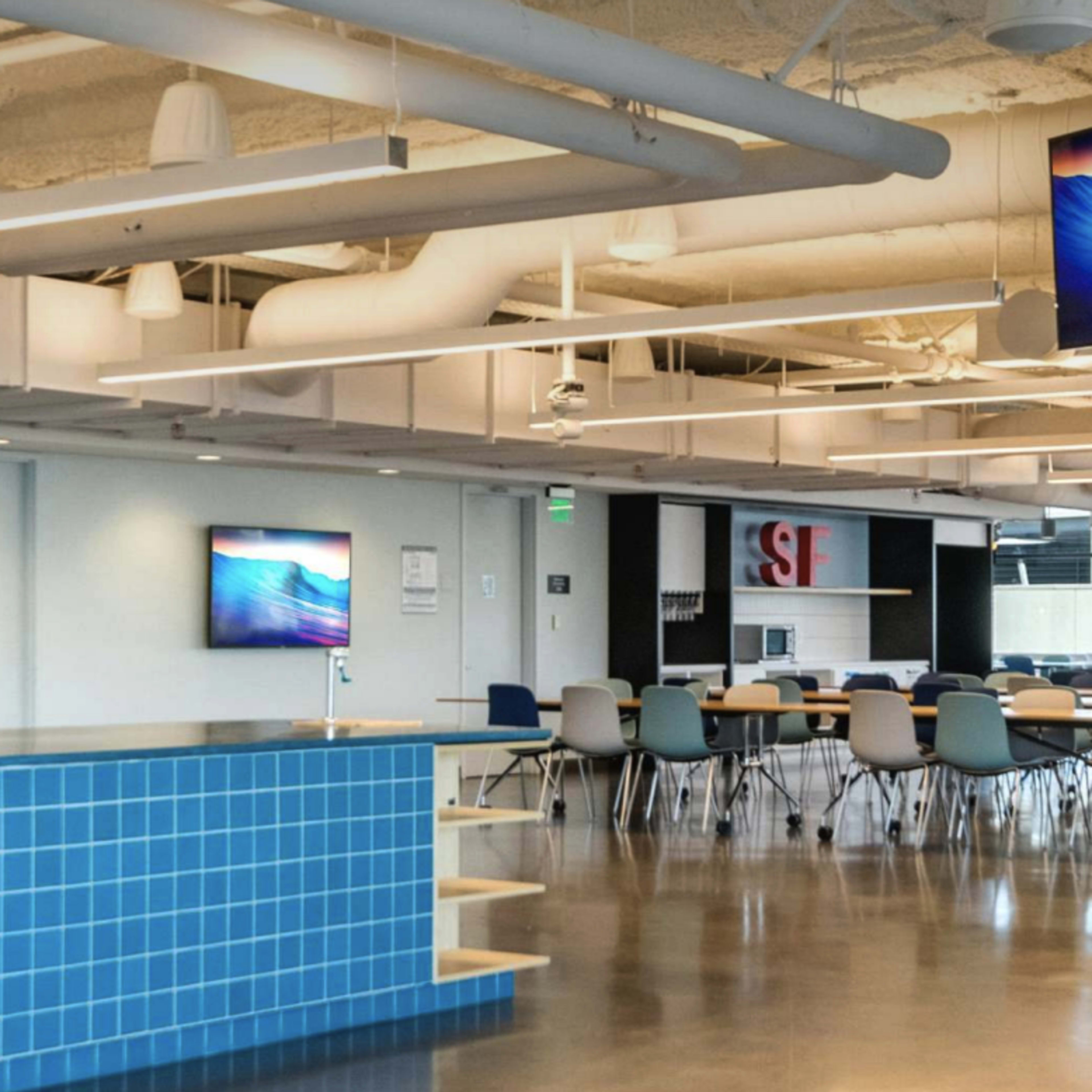 A modern office space featuring a blue tiled reception area, multiple screens displaying images, and a dining area with tables and chairs.