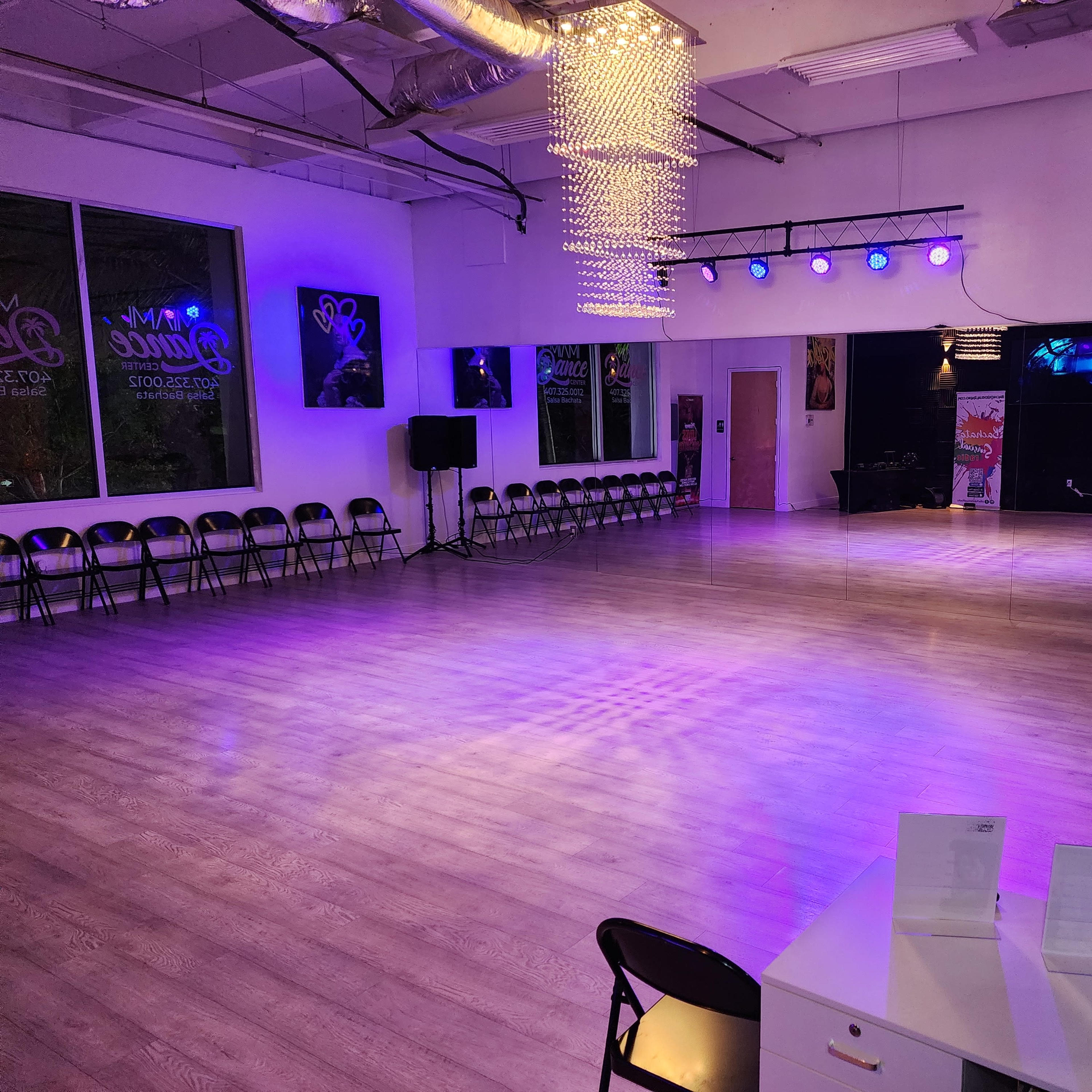 A spacious dance studio with illuminated purple lights, empty folding chairs lined against the walls, and a sound setup in the corner.