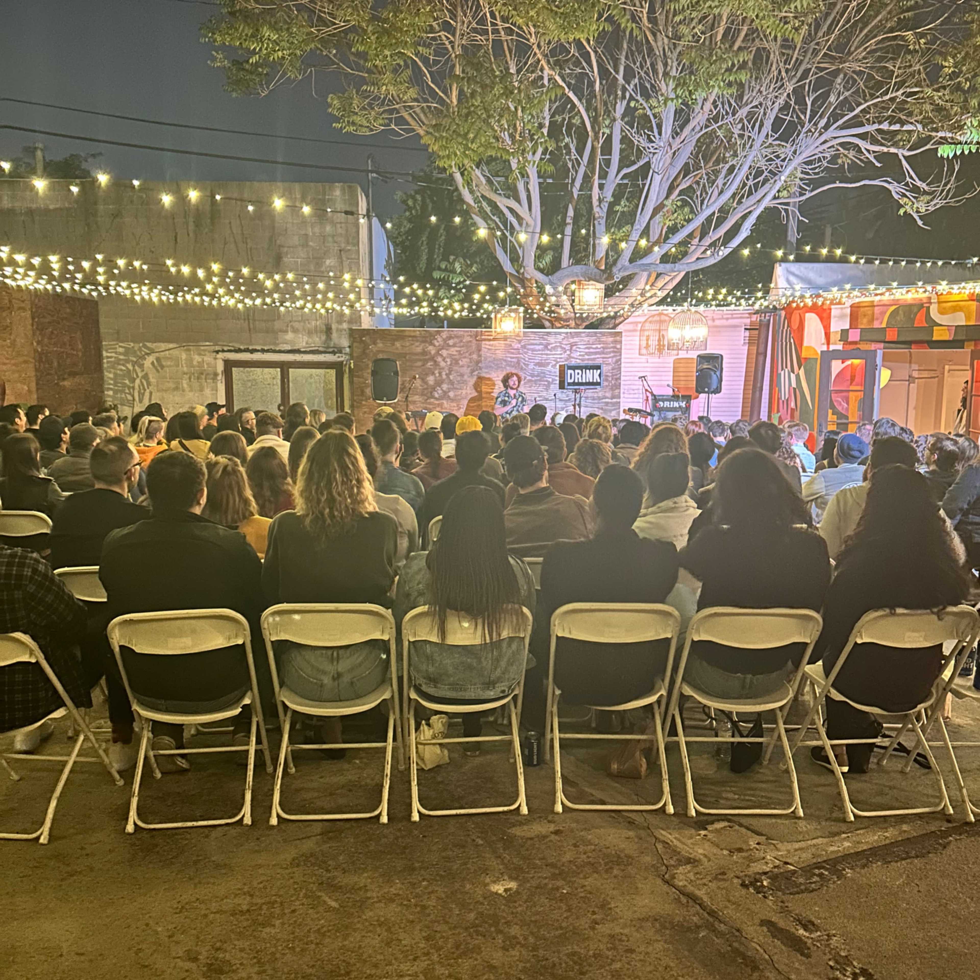 A large audience sits in folding chairs under string lights in an outdoor venue, facing a small stage.