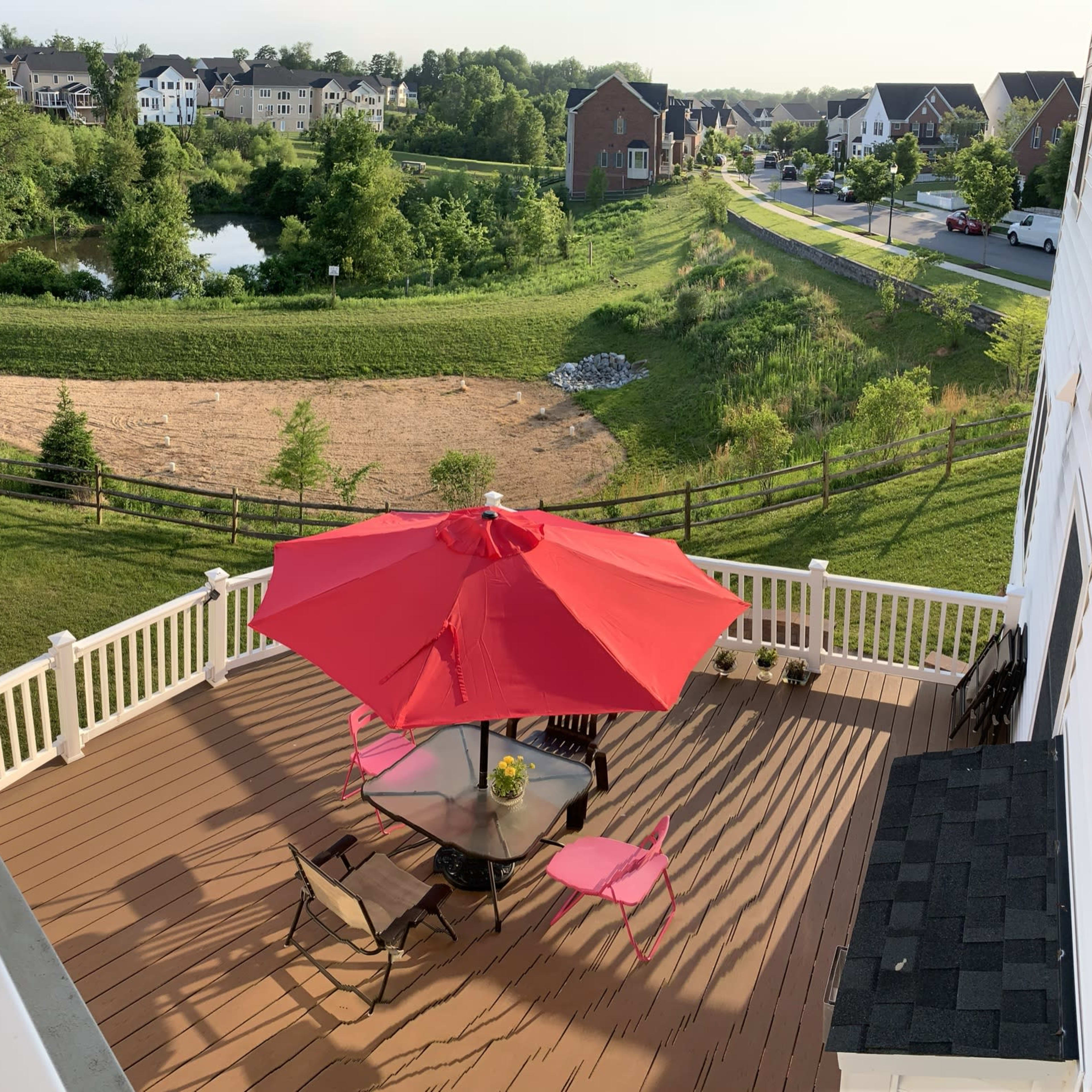A spacious deck features a round table with black chairs and a large red umbrella, overlooking a grassy area with a pond and nearby houses.