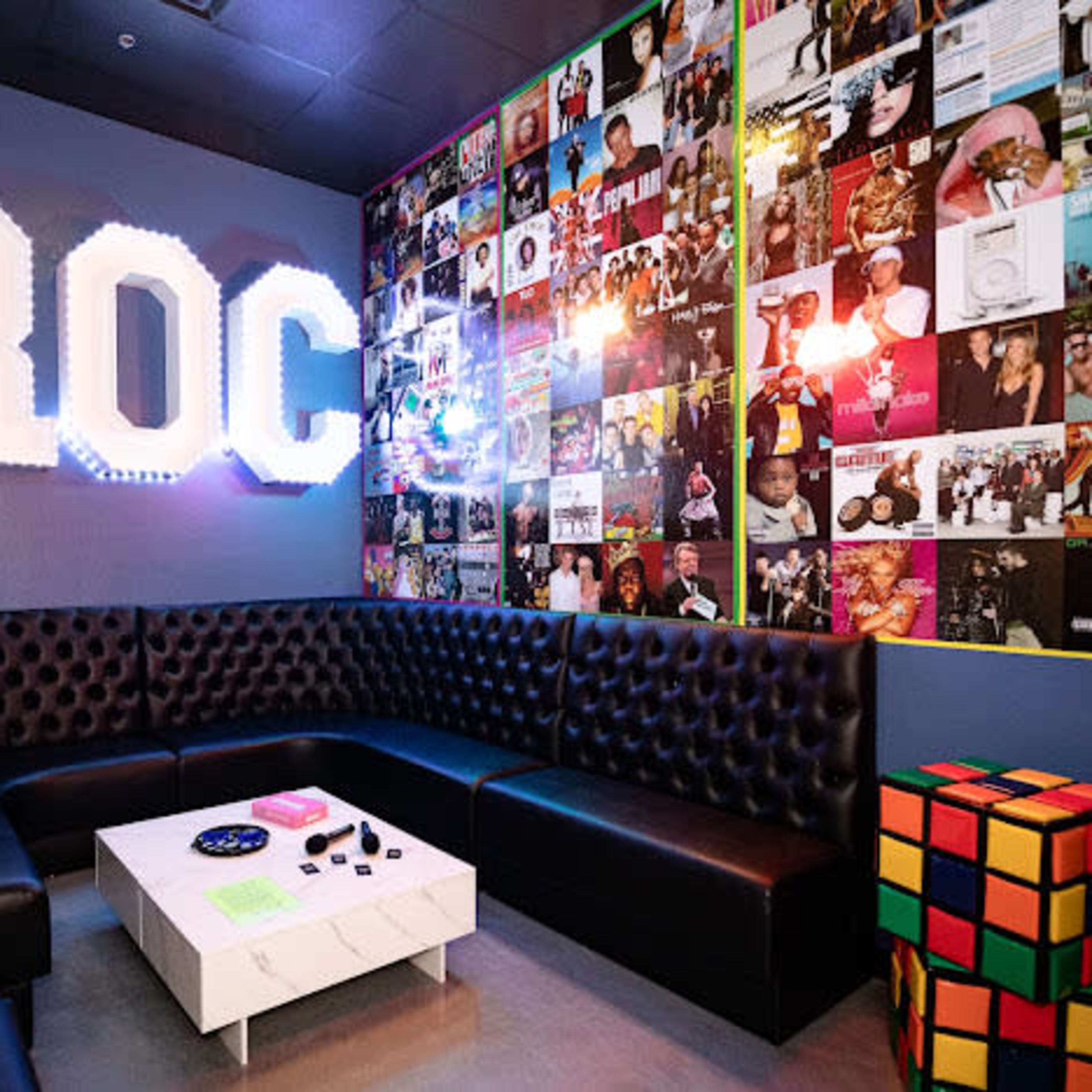 A small lounge area with a black leather sectional sofa, a white coffee table, and a wall adorned with colorful album covers.