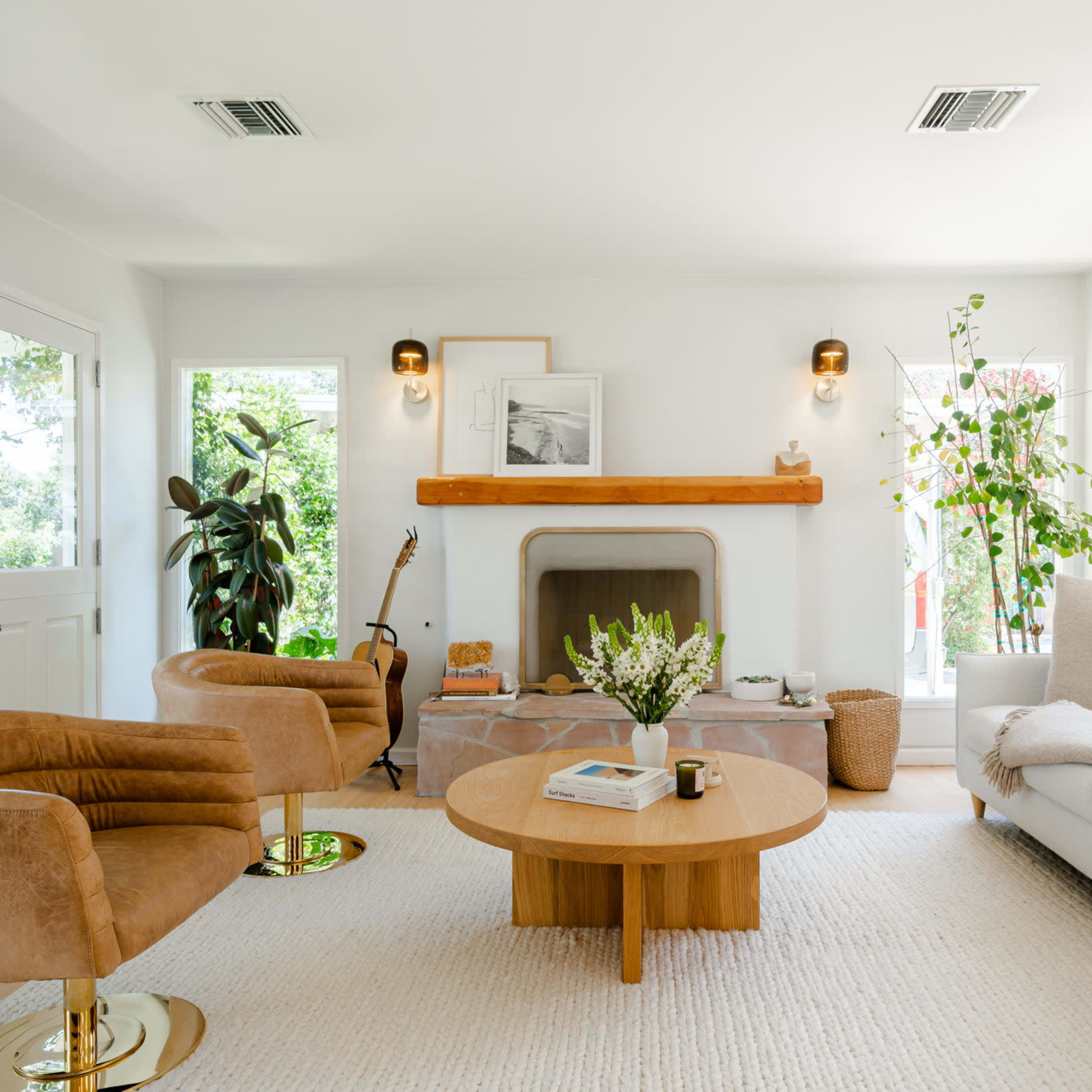The living room features a light-colored, modern design with a round wooden coffee table, two tan armchairs, and a cozy sofa arranged around a fireplace.