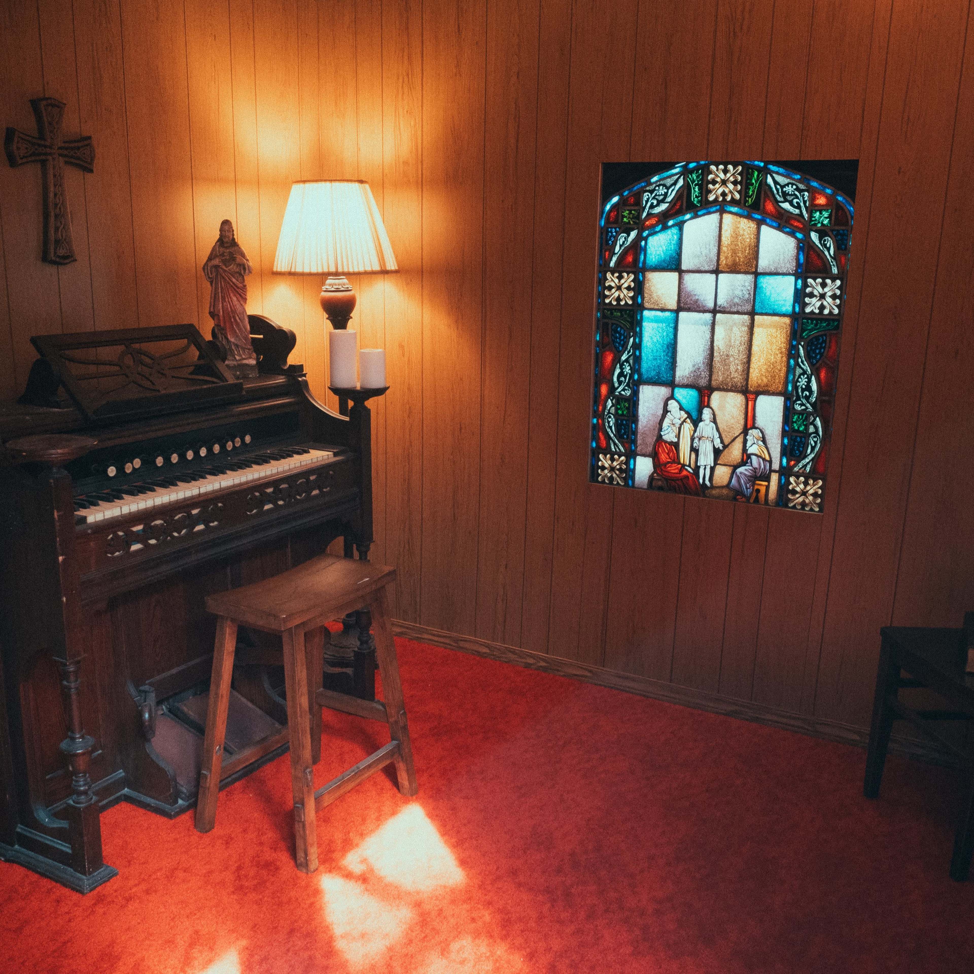 A small wooden room with a vintage organ, a lamp, a stained glass window depicting figures, and a single chair.