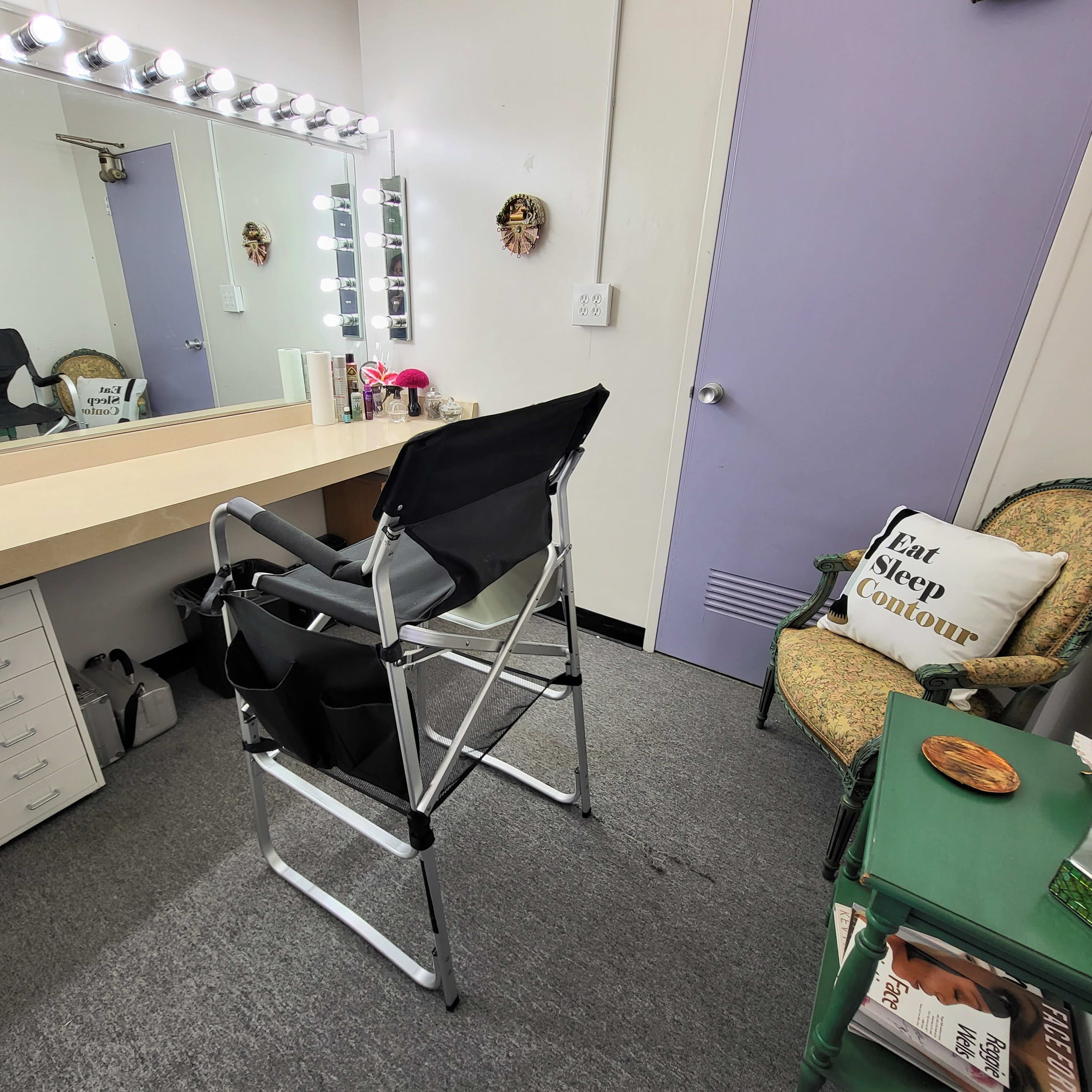 A makeup room with a large mirror, bright lights, a director's chair, and a green table holding a snack.