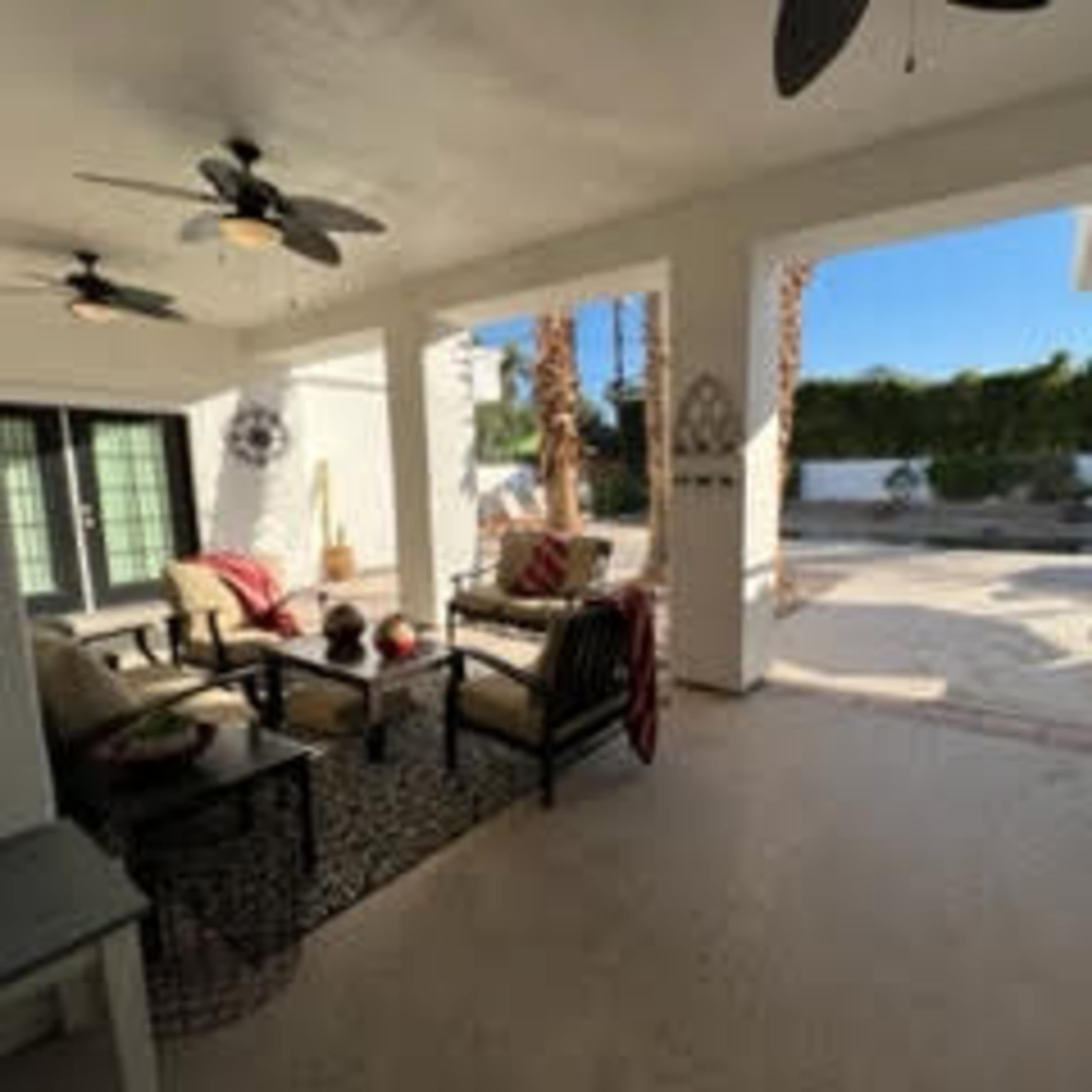 A covered patio features seating with a coffee table, surrounded by ceiling fans and a view of a fenced outdoor area.