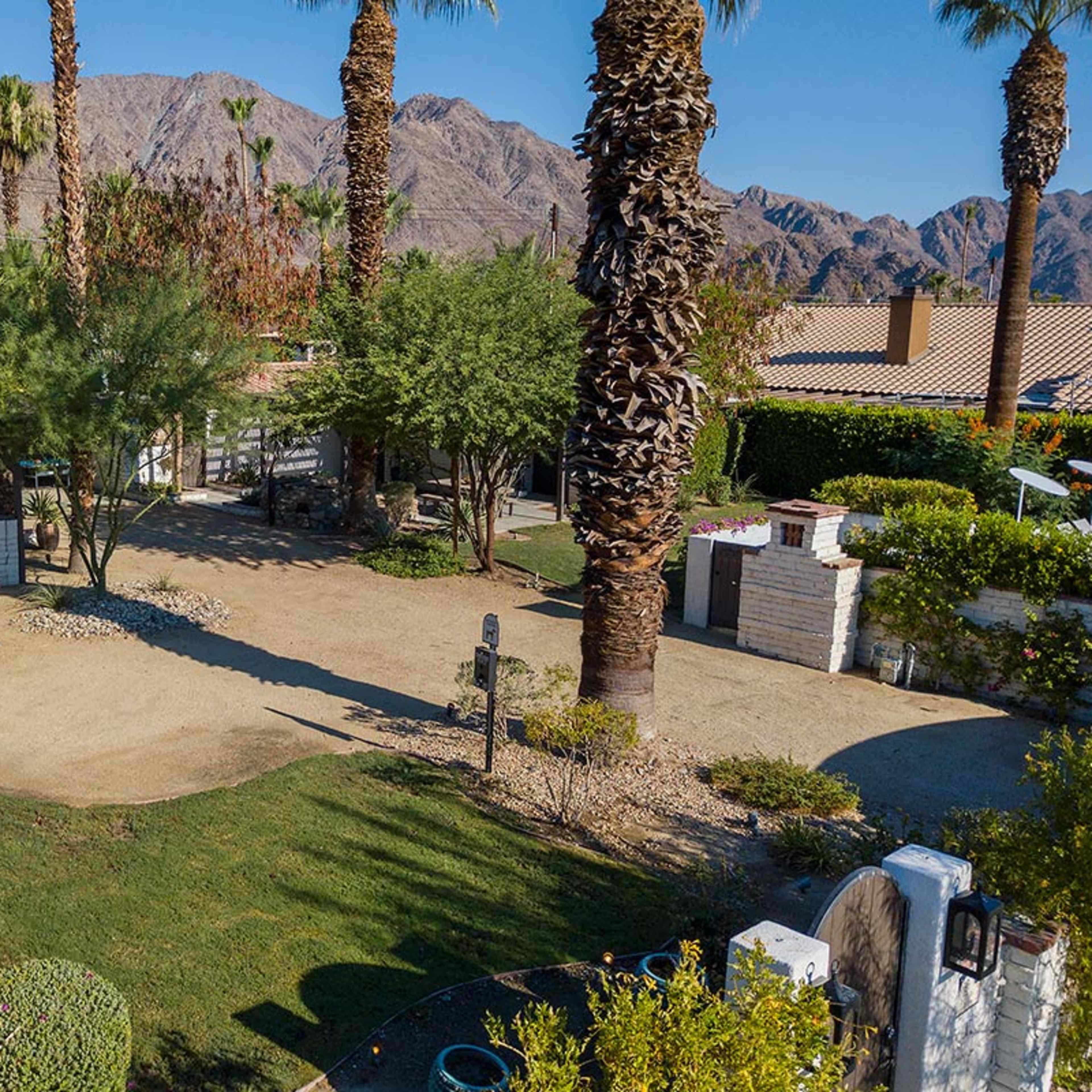 A landscaped outdoor area featuring palm trees, a gravel pathway, and a seating arrangement with white chairs near a small structure, all set against a backdrop of mountains.