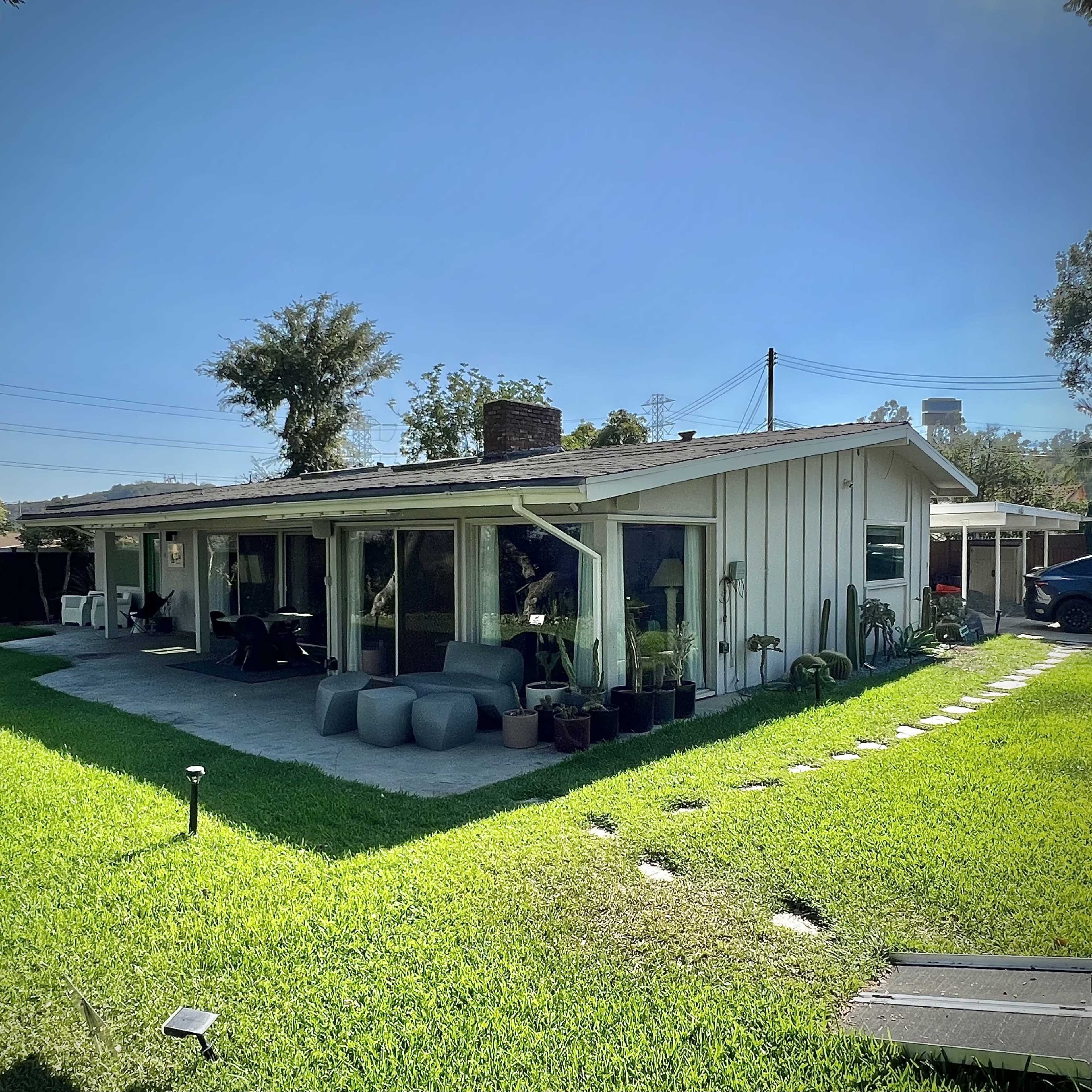 A single-story house with large windows is surrounded by a well-maintained lawn, featuring stone pathways and various potted plants.