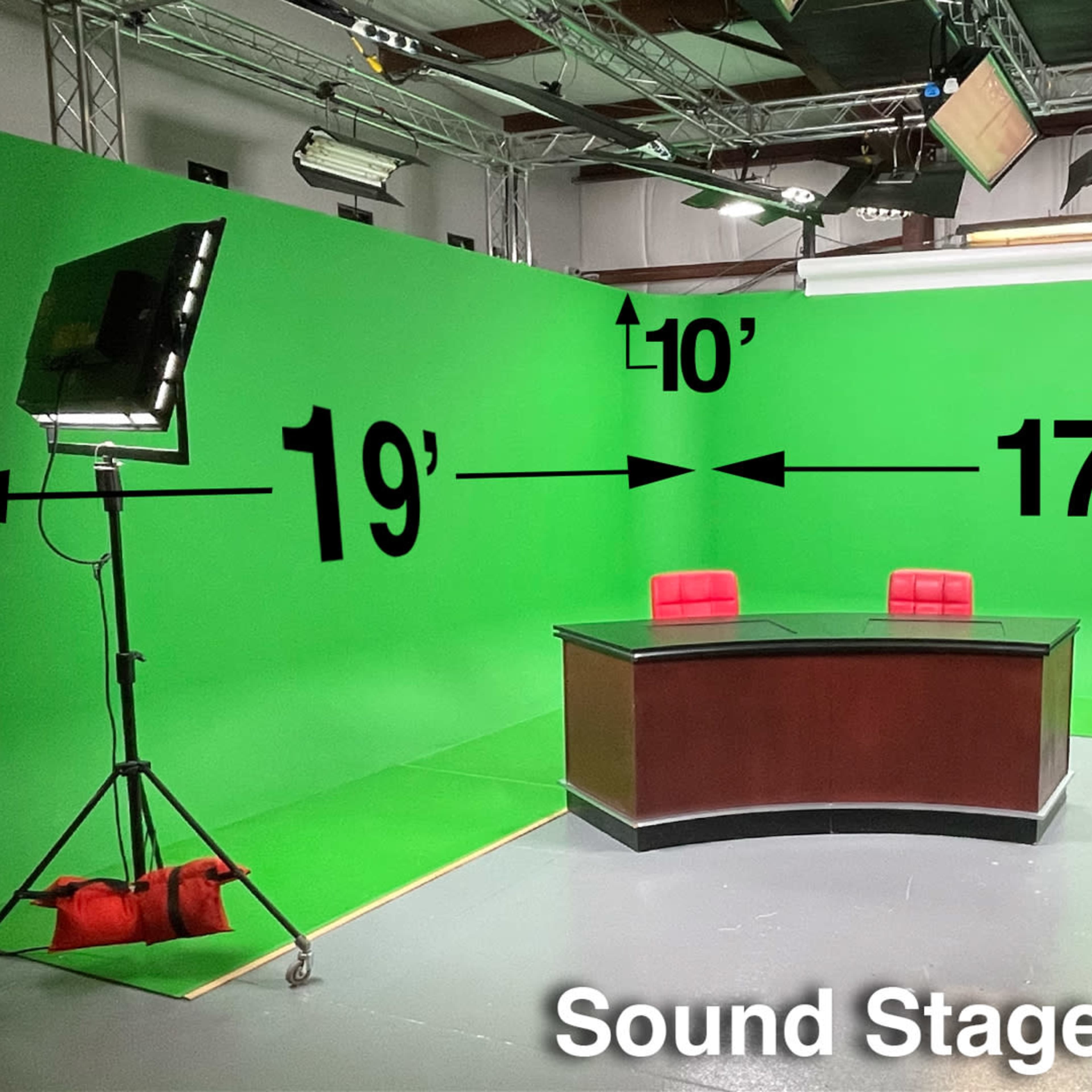 A sound stage with a green backdrop, a dark wooden desk, two red chairs, and lighting equipment set up.