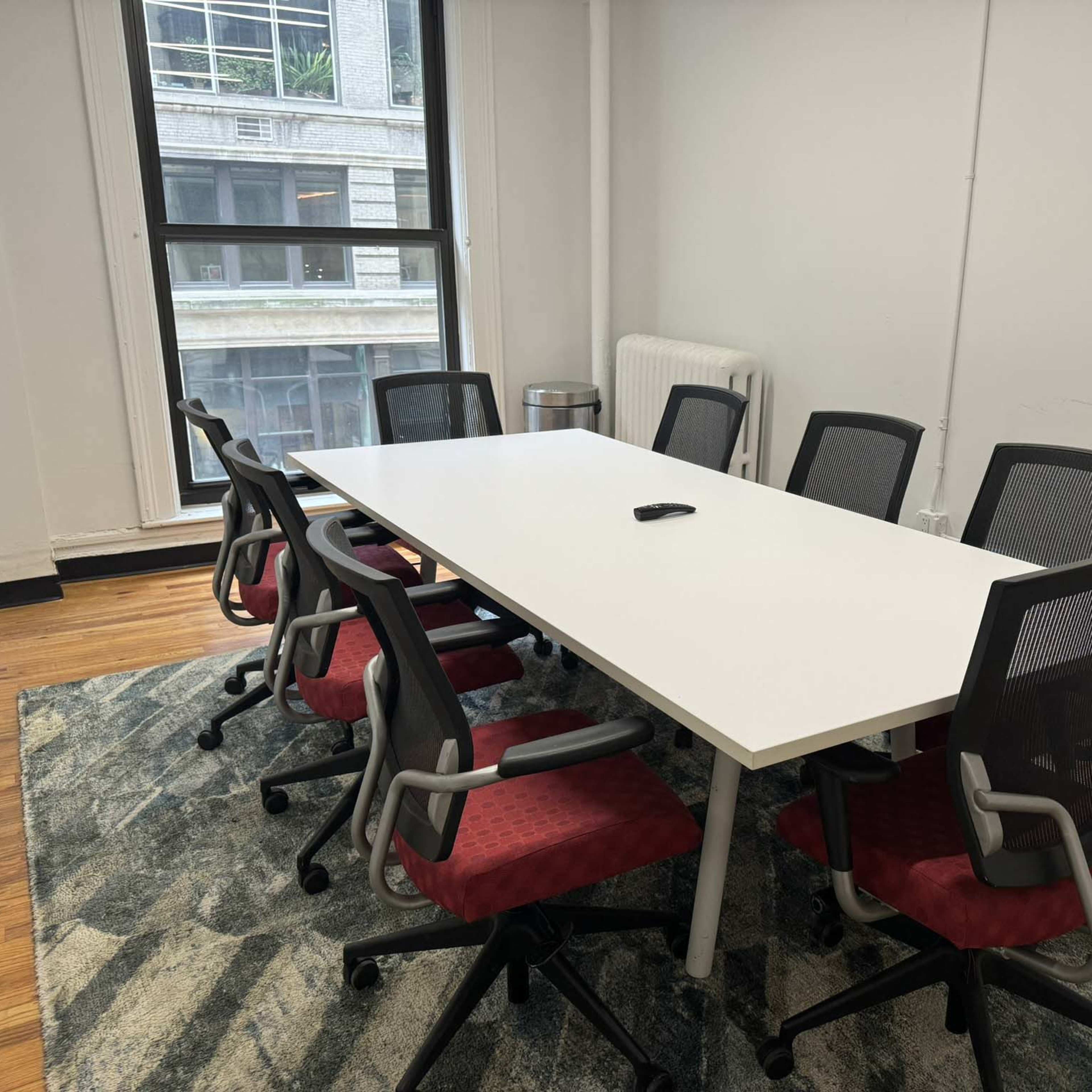 A bright conference room features a large white table surrounded by eight chairs on a patterned rug.