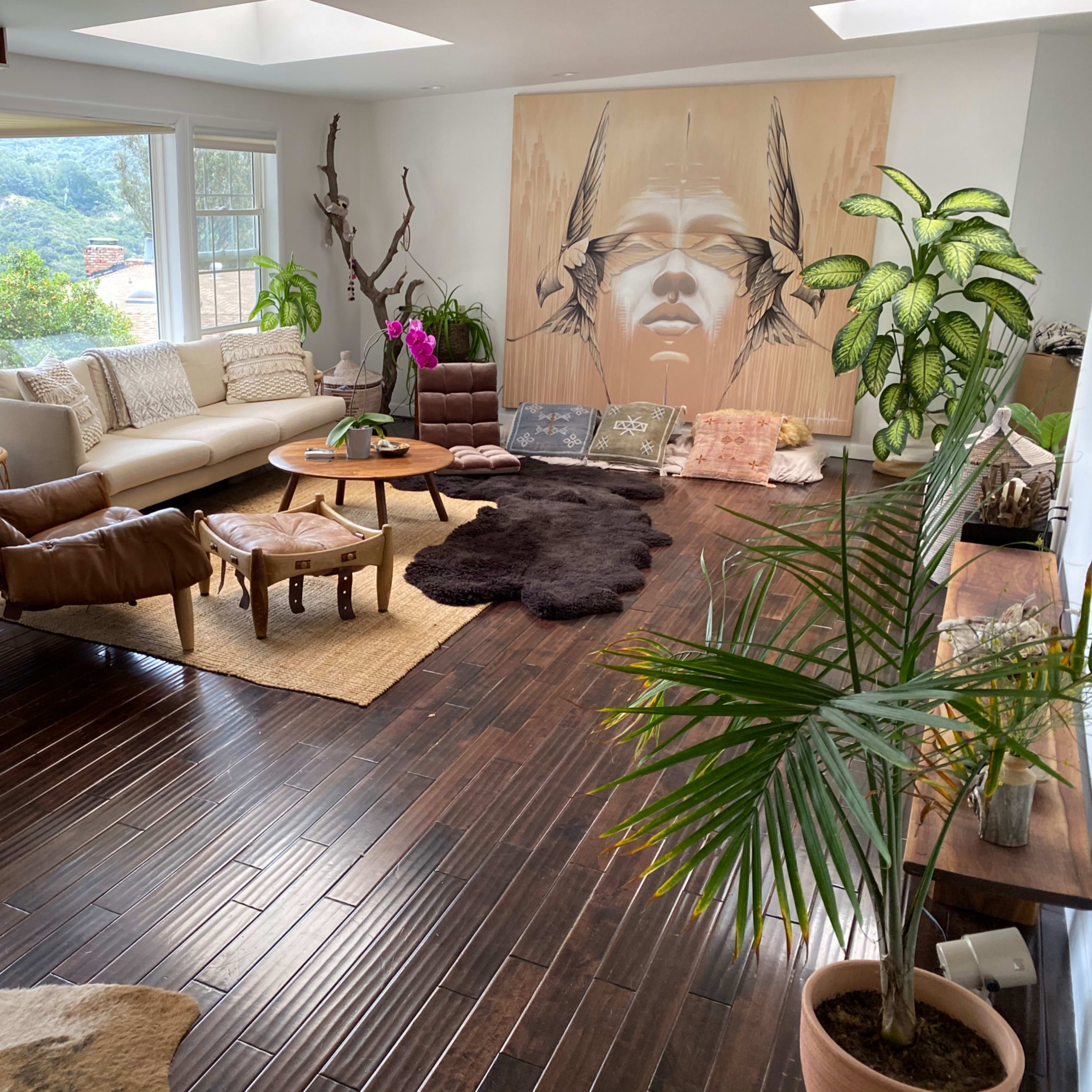 The room features a spacious sitting area with a large white sofa, leather chairs, a wooden coffee table, and an artwork depicting a face on the wall, surrounded by plants and natural light.