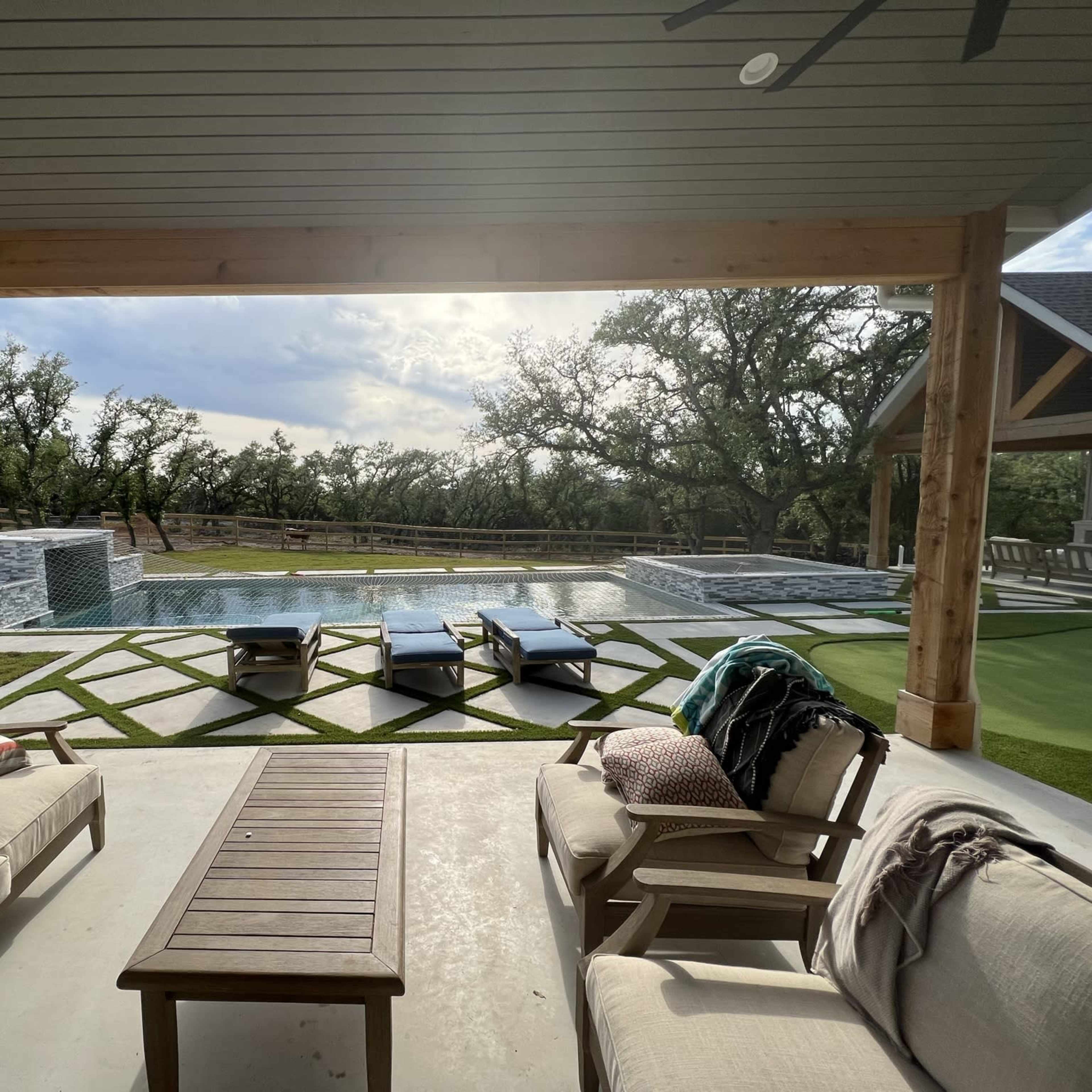A covered outdoor area features a seating arrangement facing a swimming pool bordered by landscaped greenery and a wooden fence.