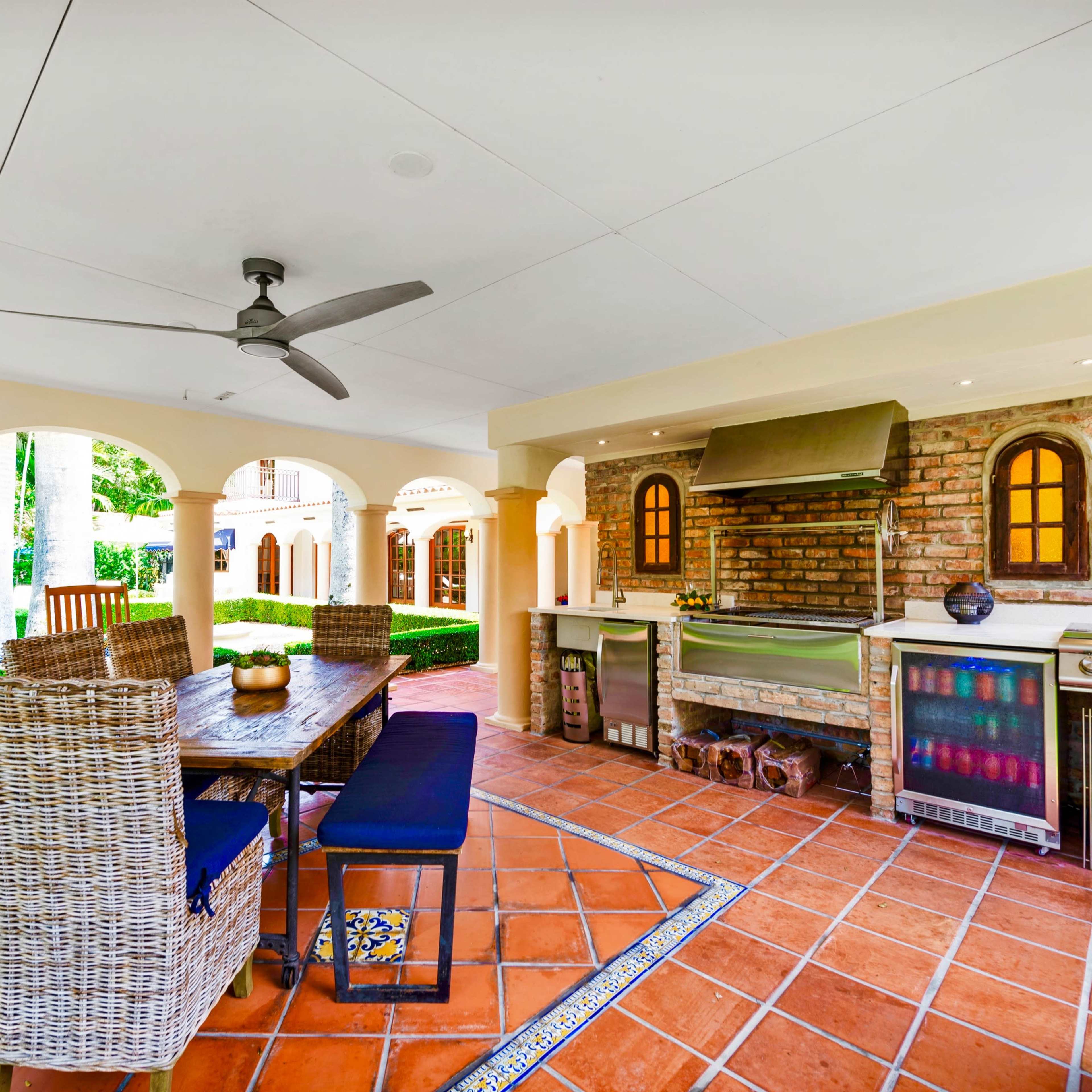 An outdoor kitchen and dining area with a brick grill, wooden dining table, and wicker chairs under a covered patio.