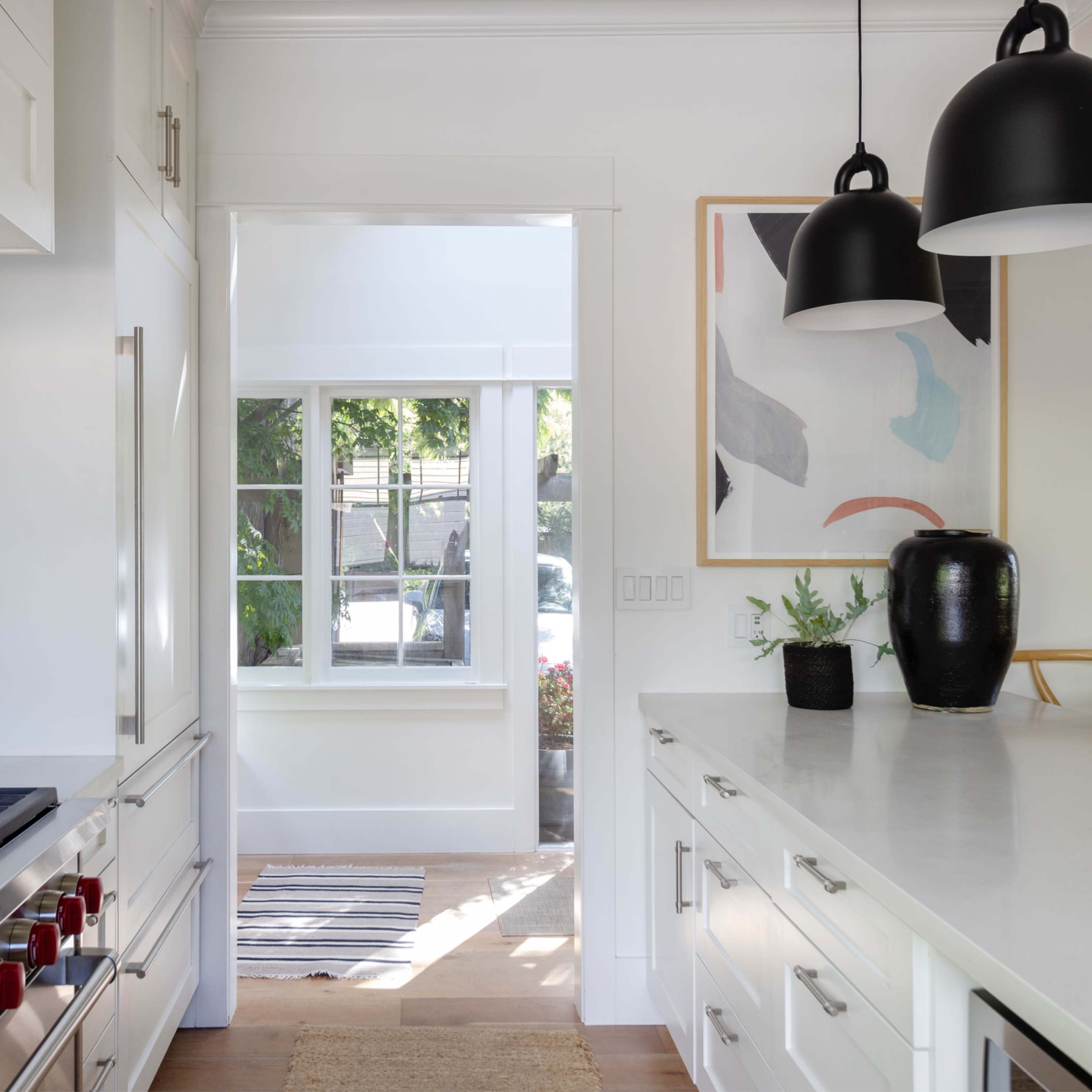 A modern kitchen with white cabinetry, a large gas stove, and a view of a hallway leading to a well-lit area with an abstract painting and plants.