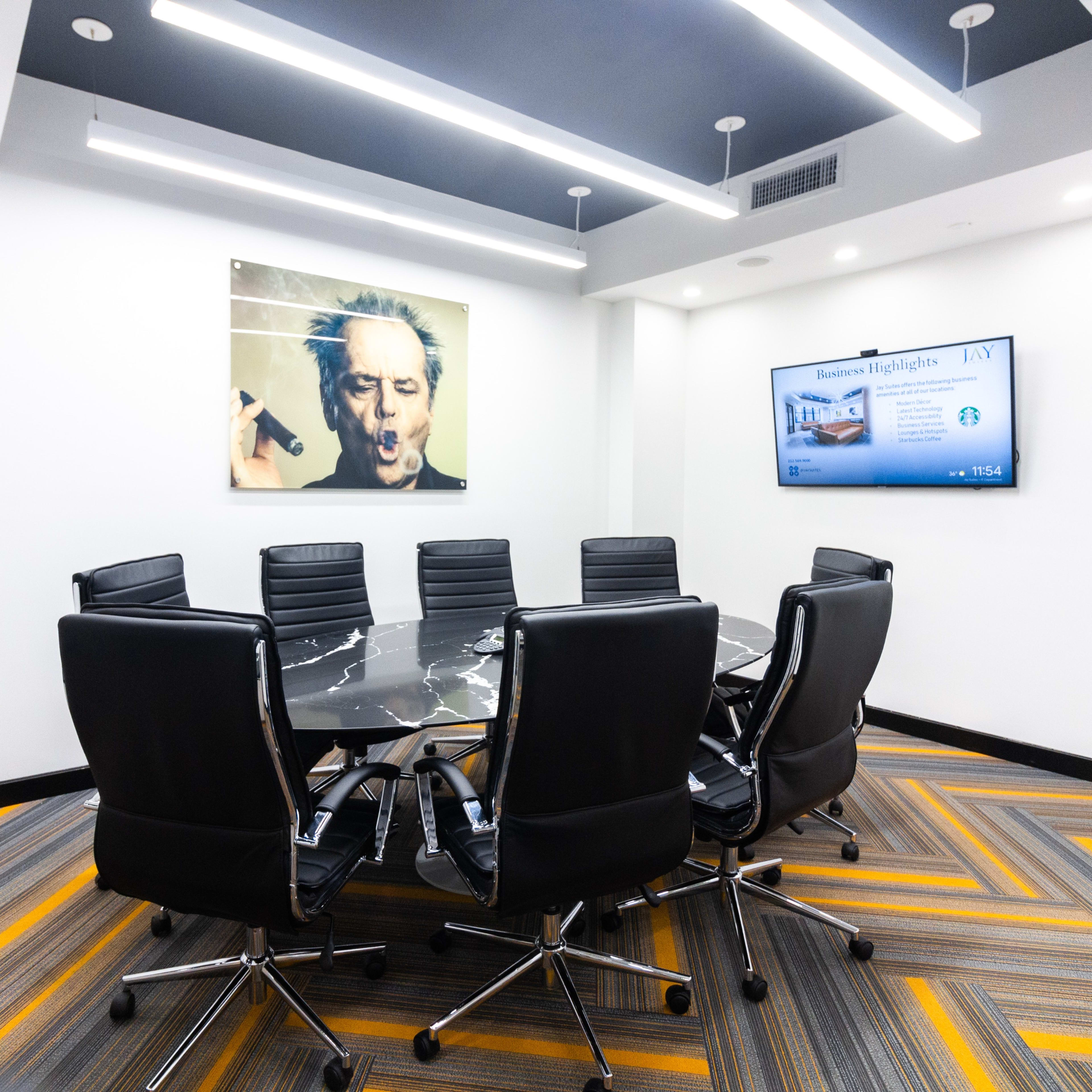 A modern conference room features a large black marble table surrounded by eight leather chairs, with an artwork and a screen displaying presentations on the walls.