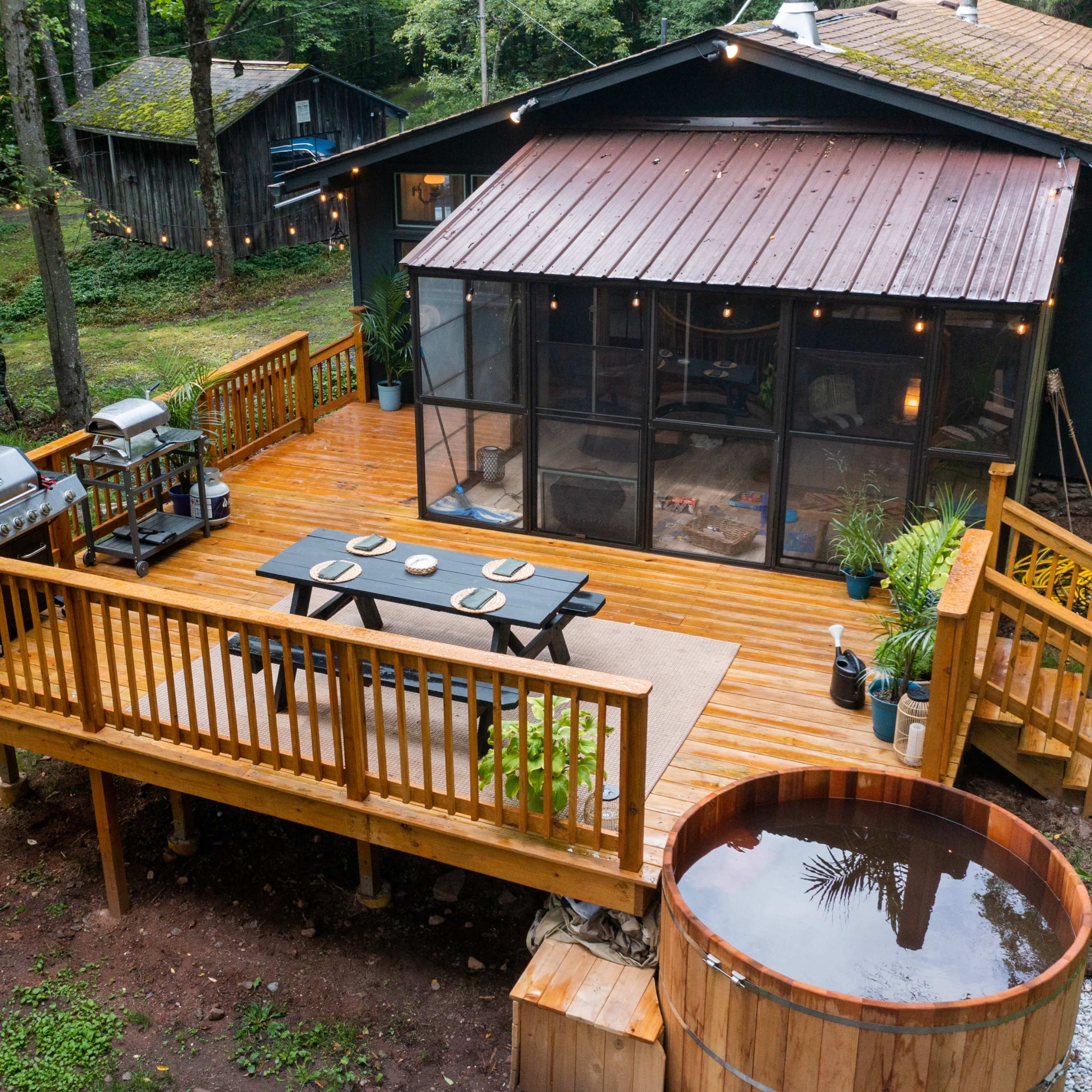 A wooden deck with a dining table and chairs is attached to a house, featuring a hot tub and grill, surrounded by trees and outdoor seating.