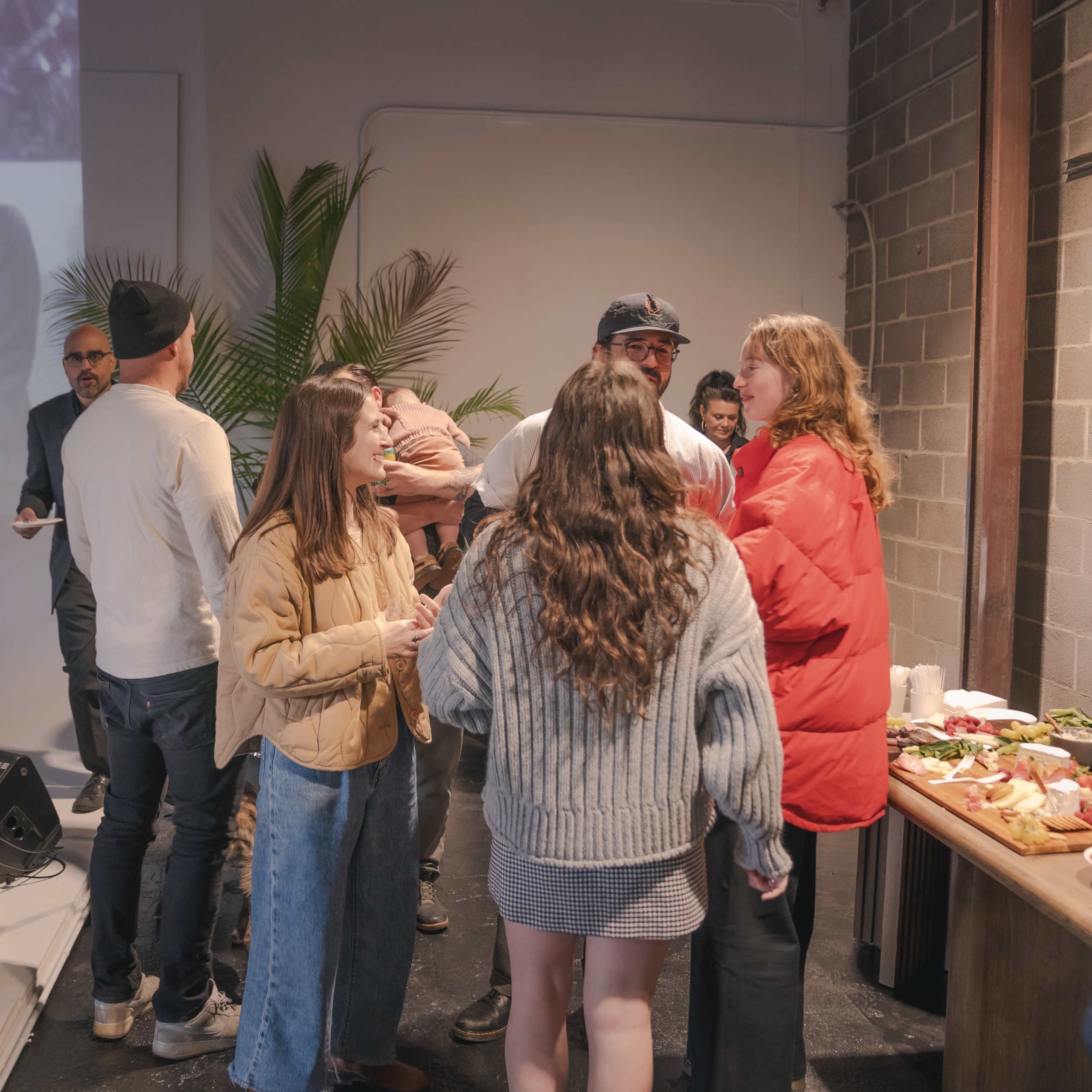 A group of people are gathered in a modern event space, engaged in conversation near a buffet table, with a projection of a black-and-white image in the background.