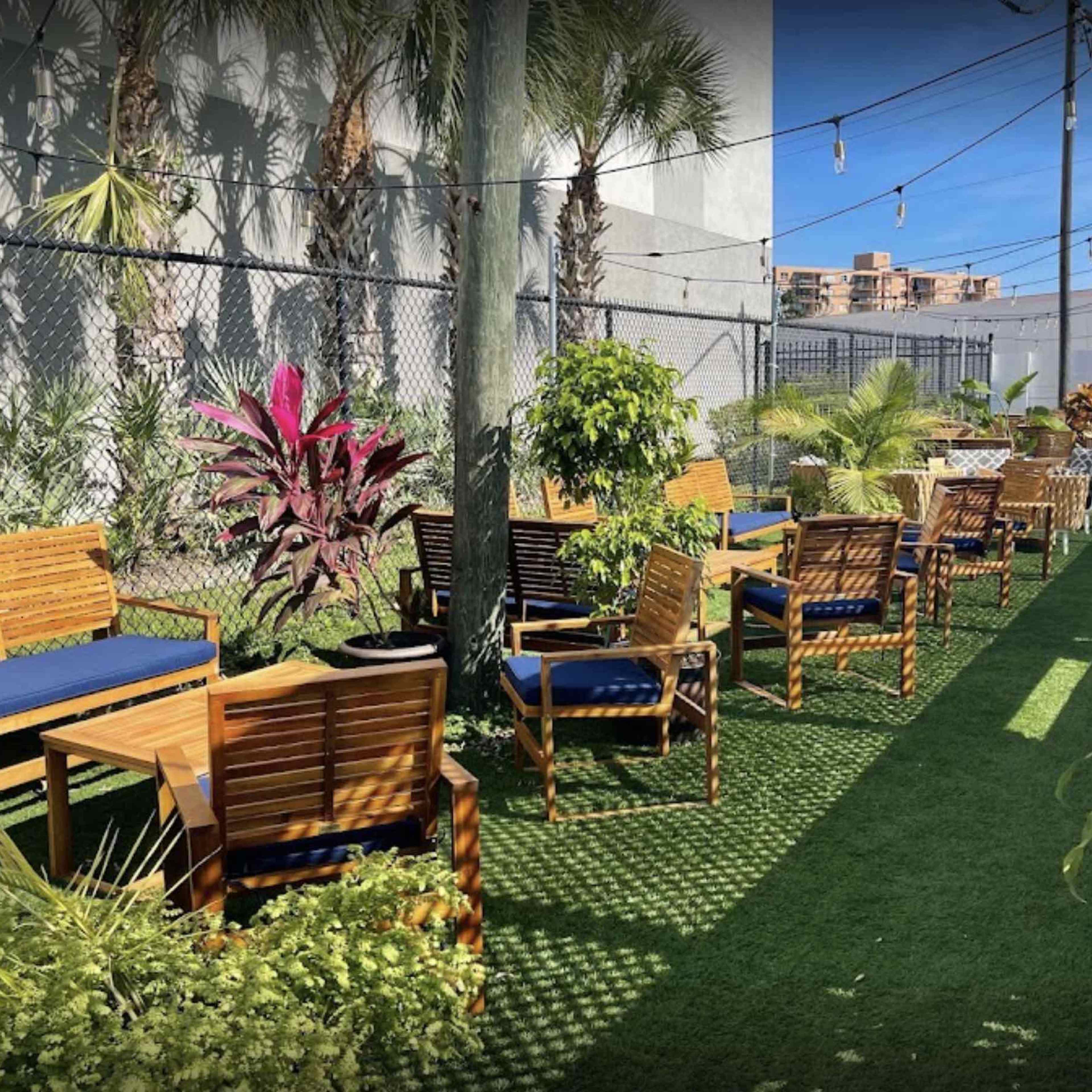 A green outdoor seating area with wooden benches and tables, surrounded by plants and a fence.