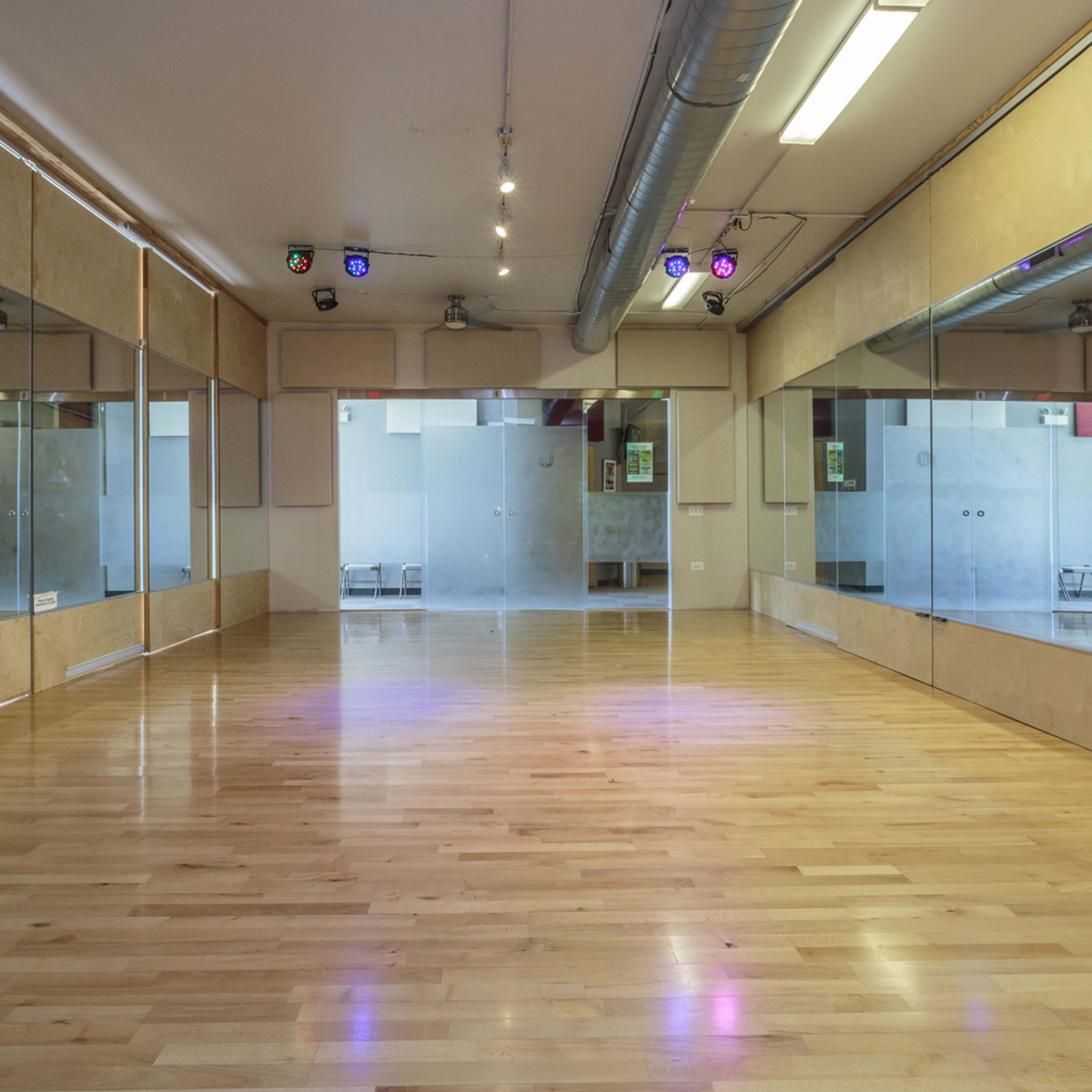 A spacious dance studio with wooden flooring, mirrored walls, and overhead lighting.