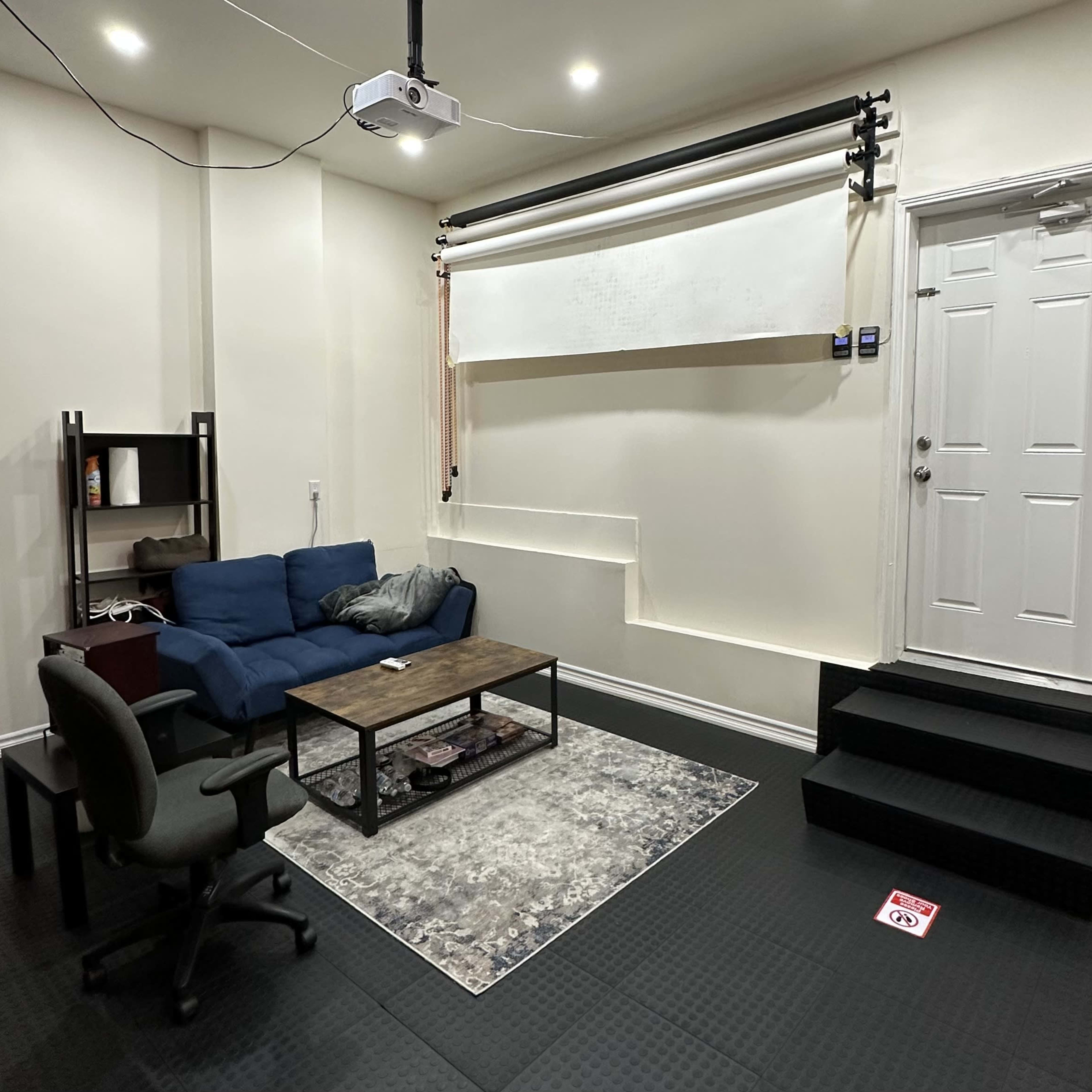 A small room with a blue couch, a wooden coffee table, an office chair, a door, and a projection screen mounted on the wall.