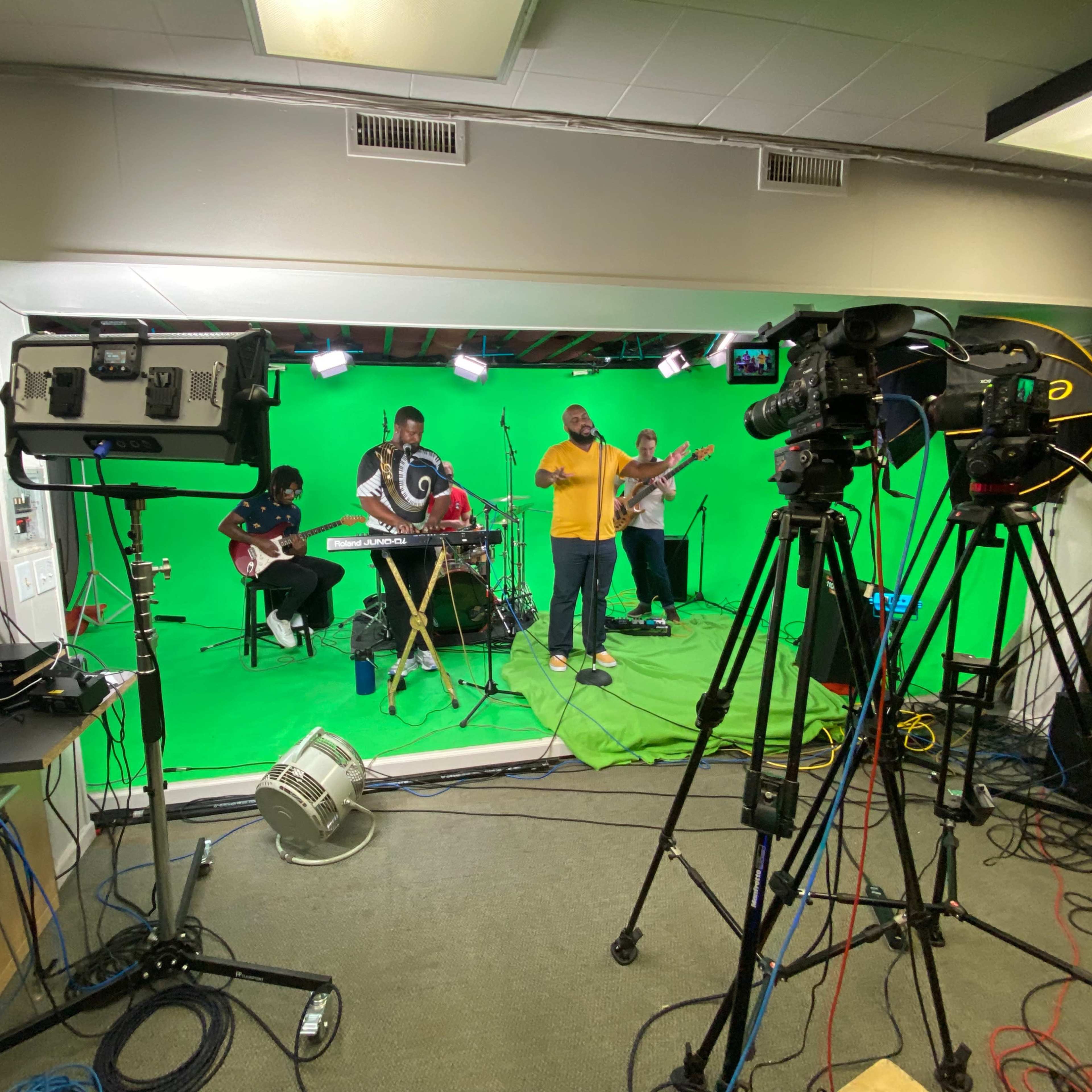 A band performs in front of a green screen, surrounded by cameras and studio equipment.