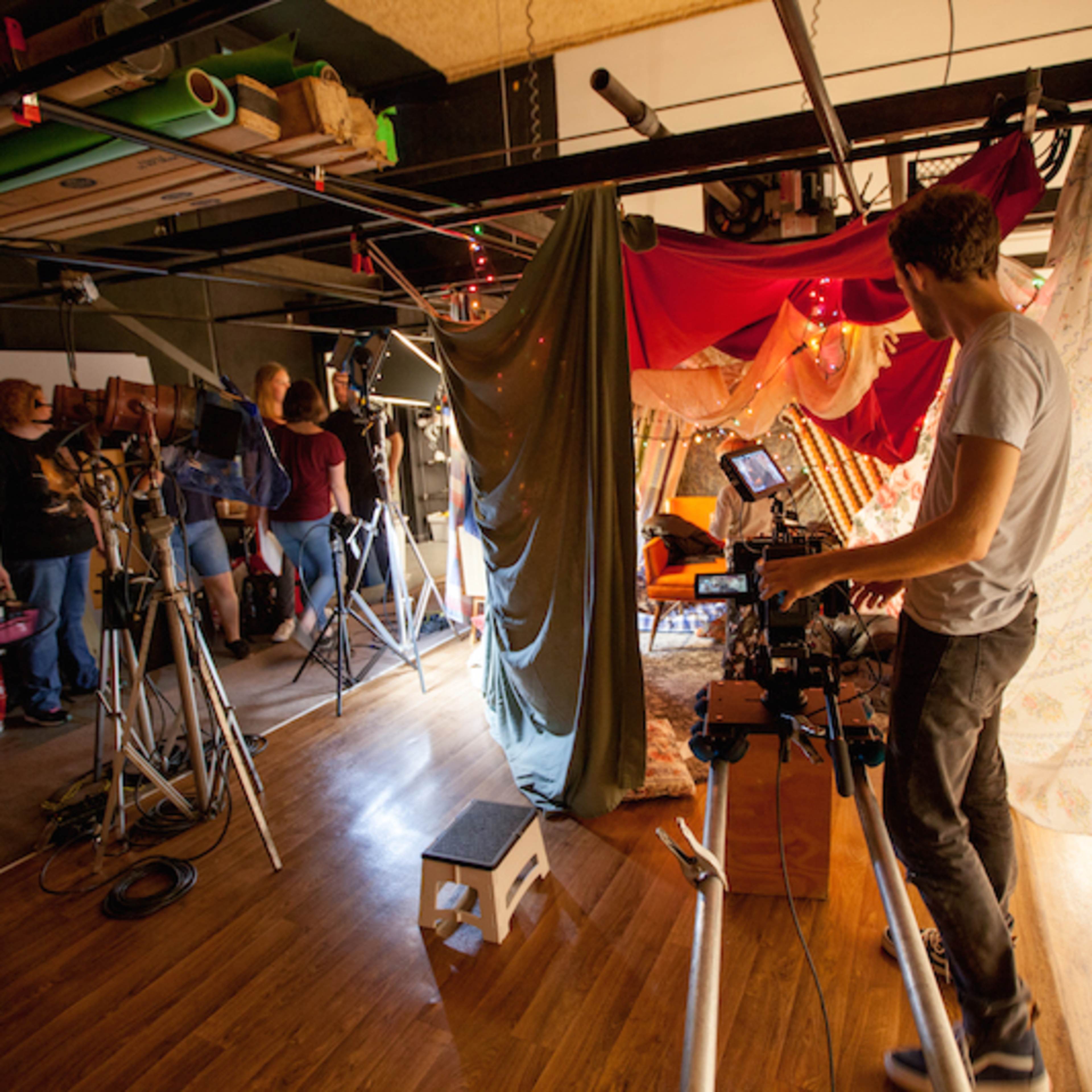 A film crew sets up equipment in a studio covered with colorful fabric drapes.