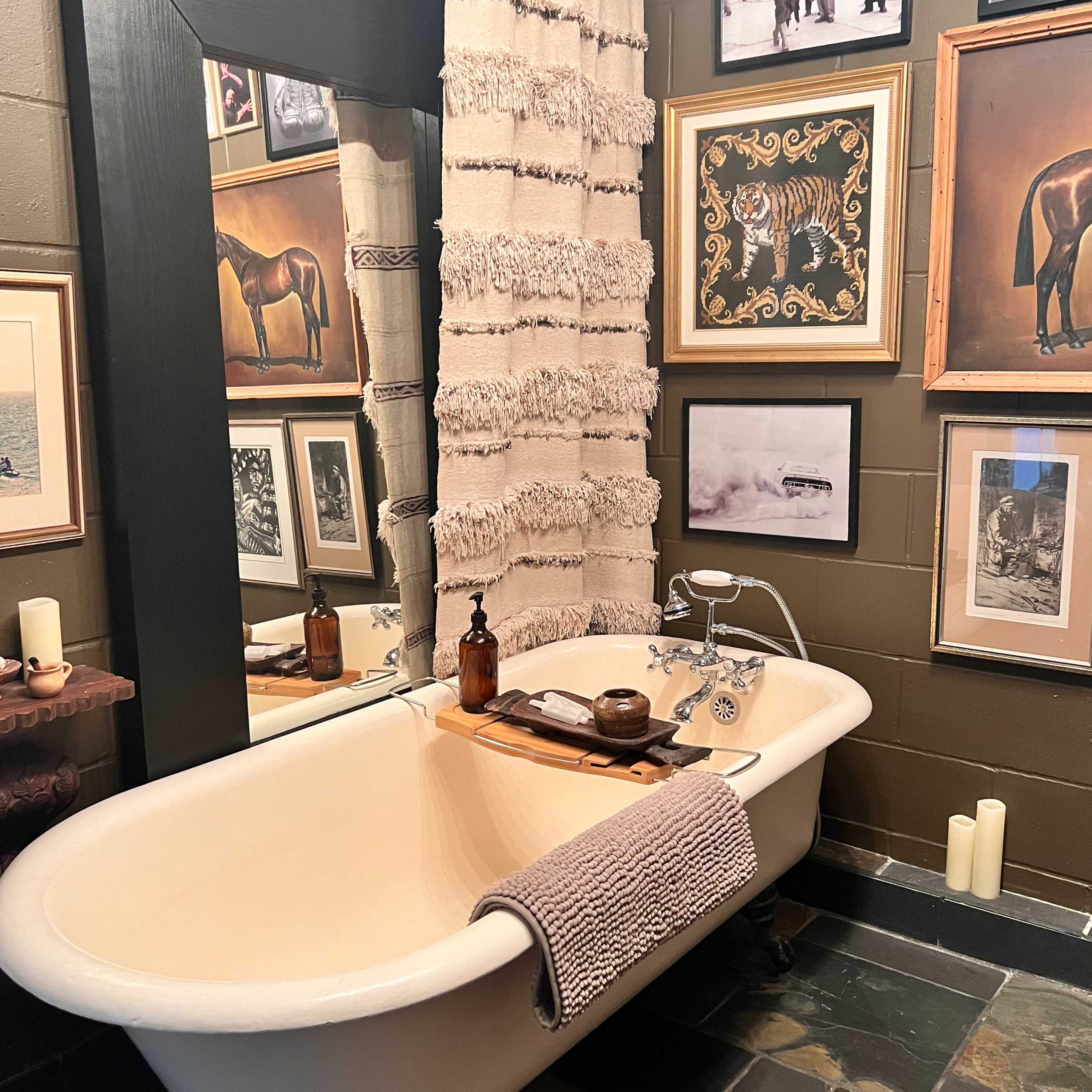 A vintage freestanding bathtub with a decorative tray on it, surrounded by various framed pictures and a textured curtain in a warmly lit bathroom.