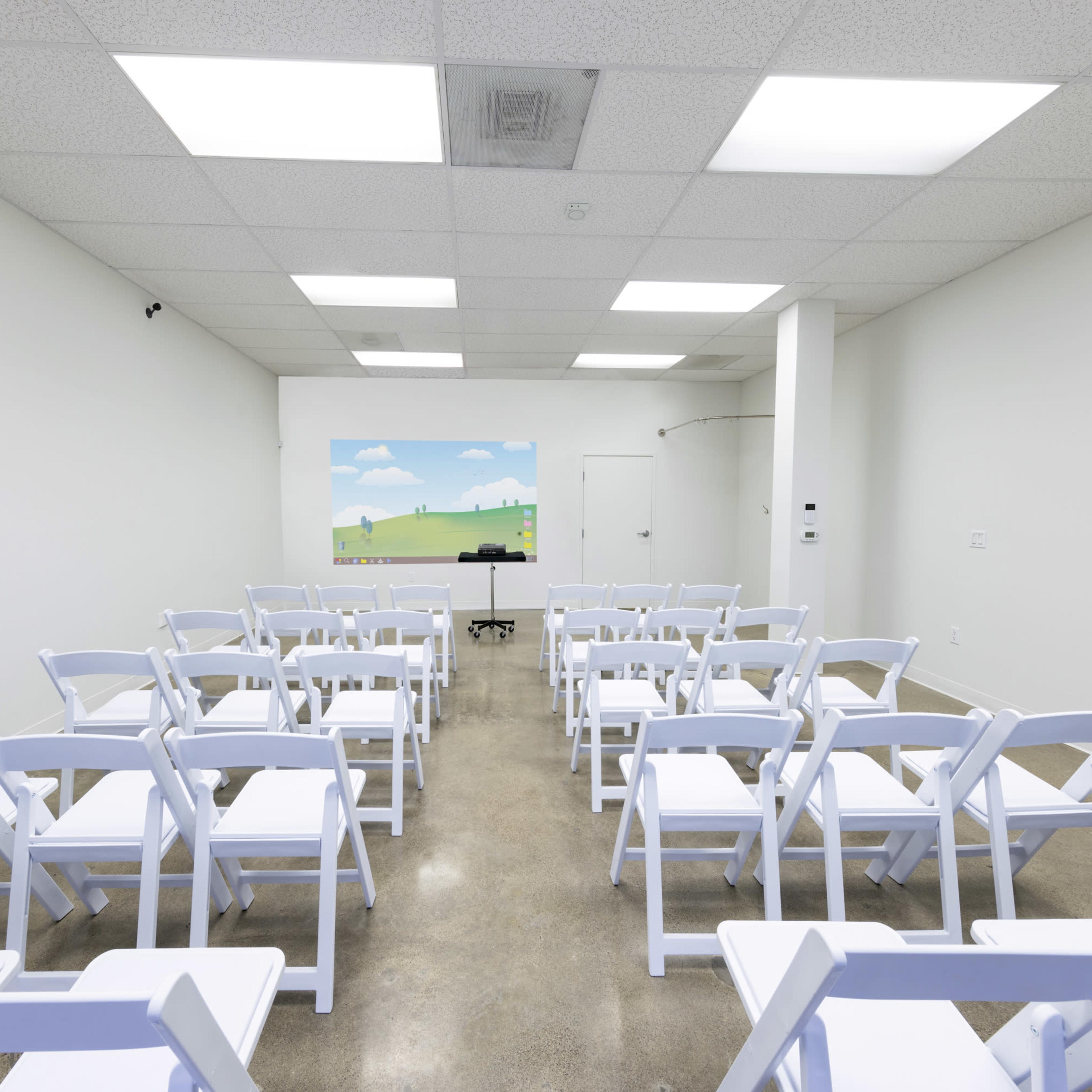 A bright, empty room features white folding chairs arranged in rows facing a screen displaying a landscape.