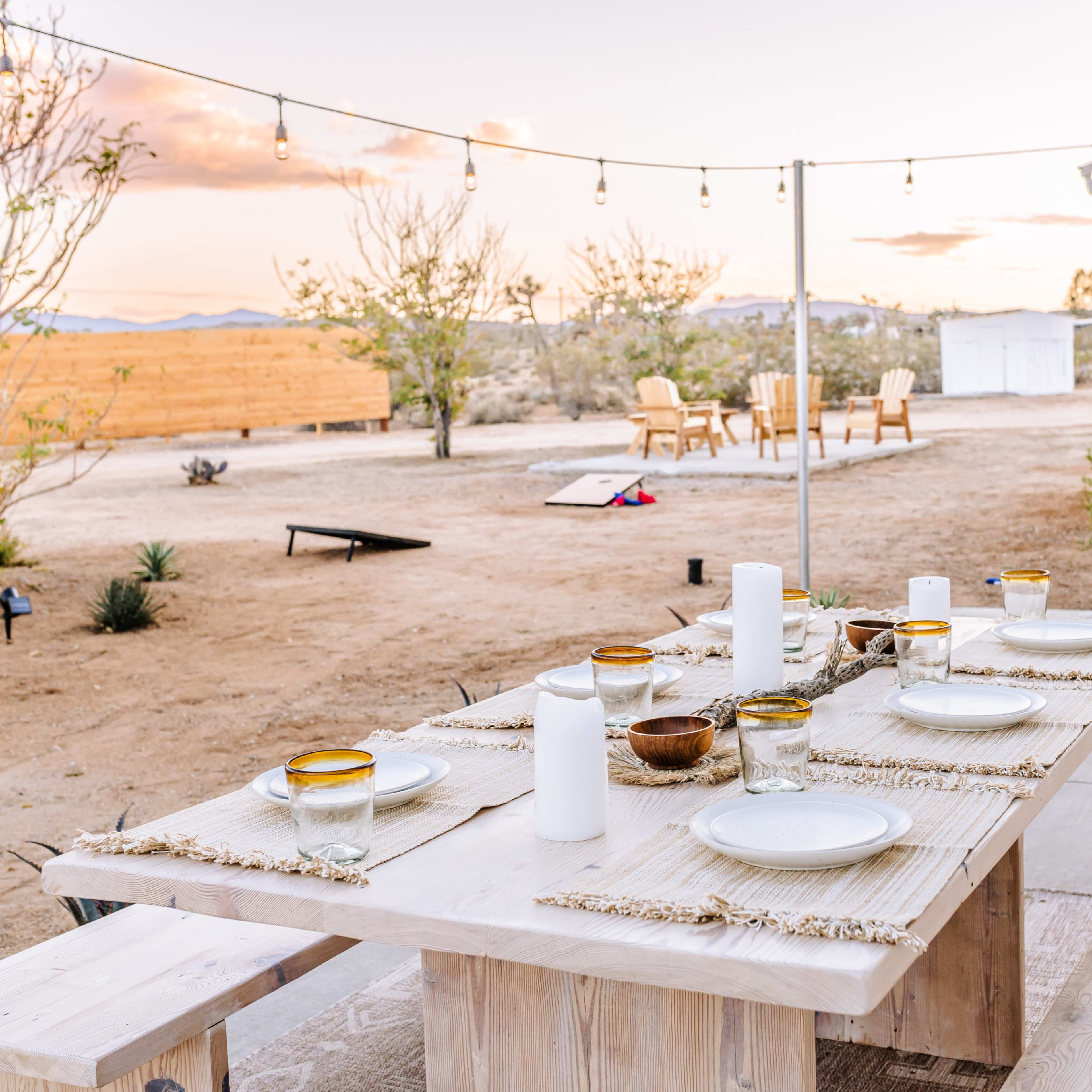 A wooden dining table is set for a meal outdoors, surrounded by desert landscaping and string lights overhead, with two chairs visible in the background.