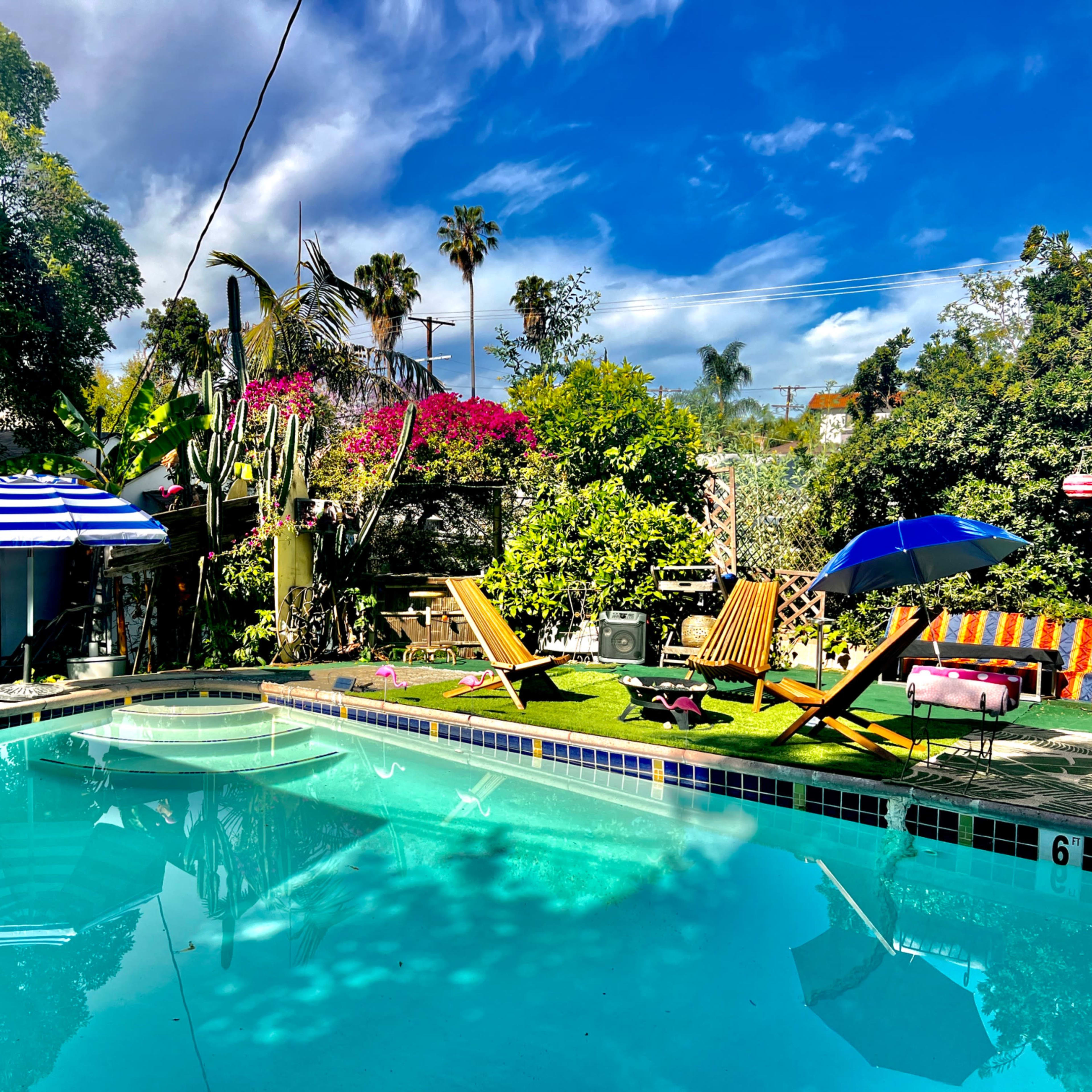 A vibrant outdoor pool area surrounded by lush greenery, with lounge chairs and umbrellas placed around the water.