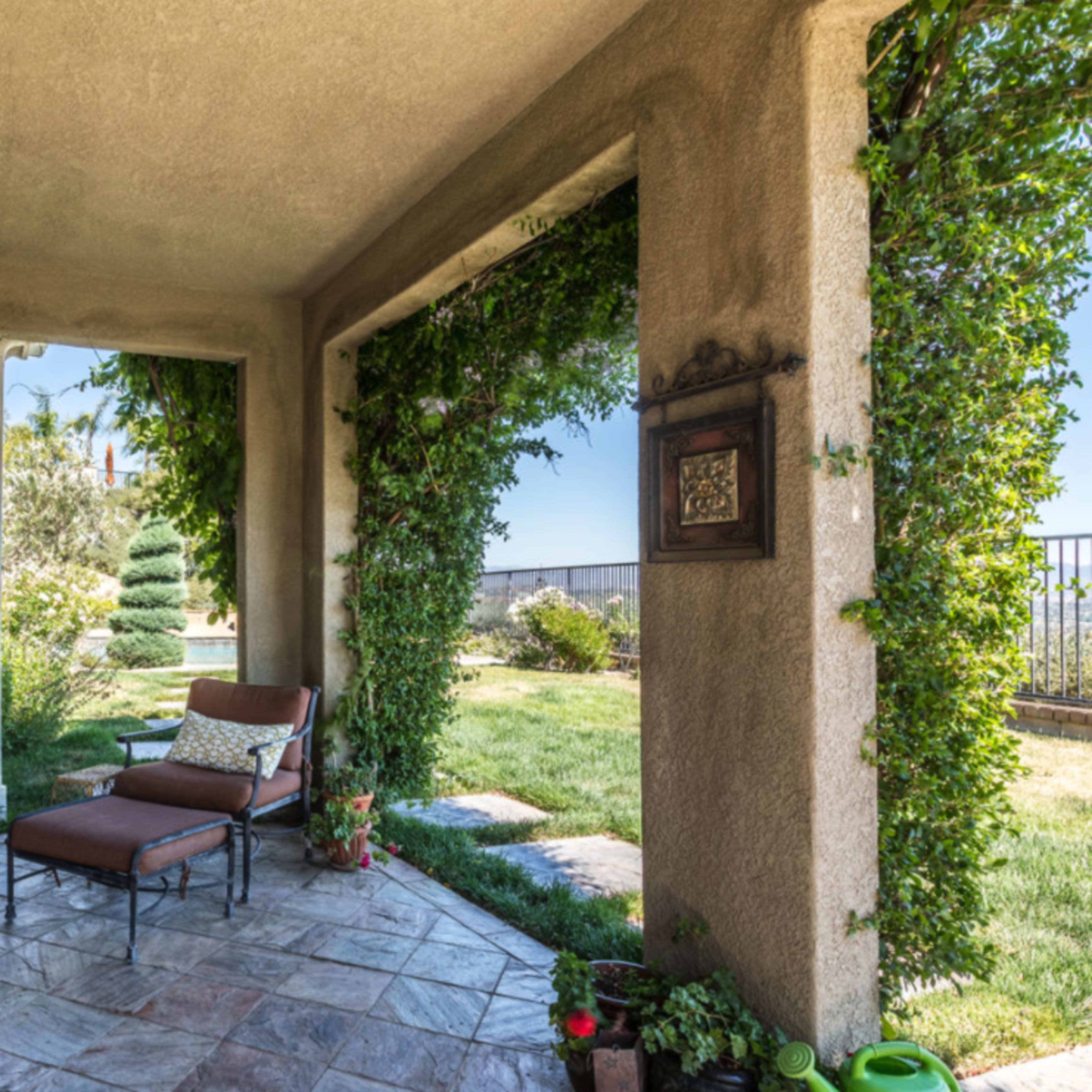 A covered patio features a lounge chair beside a potted plant, with greenery climbing the columns and a view of the lawn beyond.