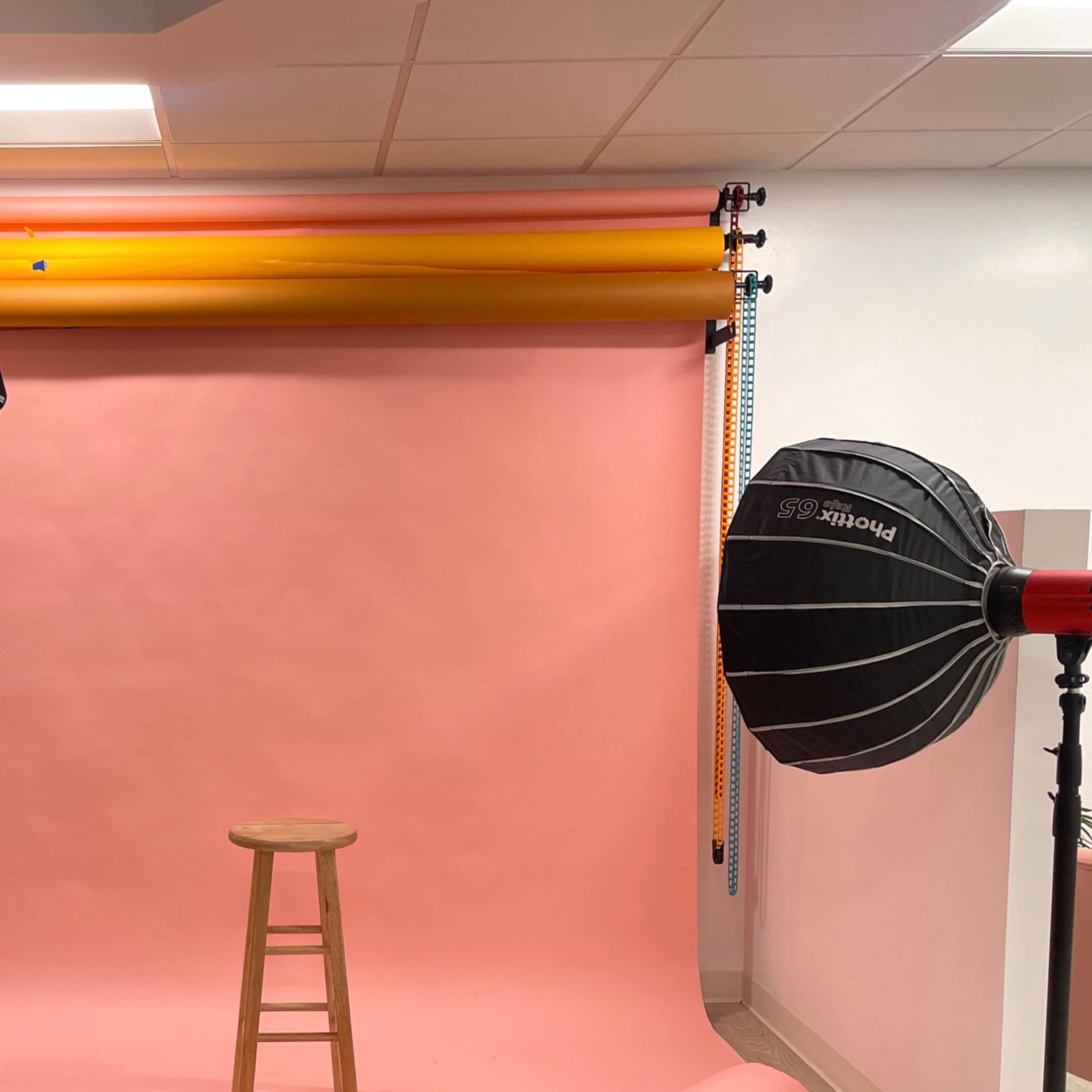 A photography studio setup with two large softboxes, a pink backdrop, and a wooden stool in the center.