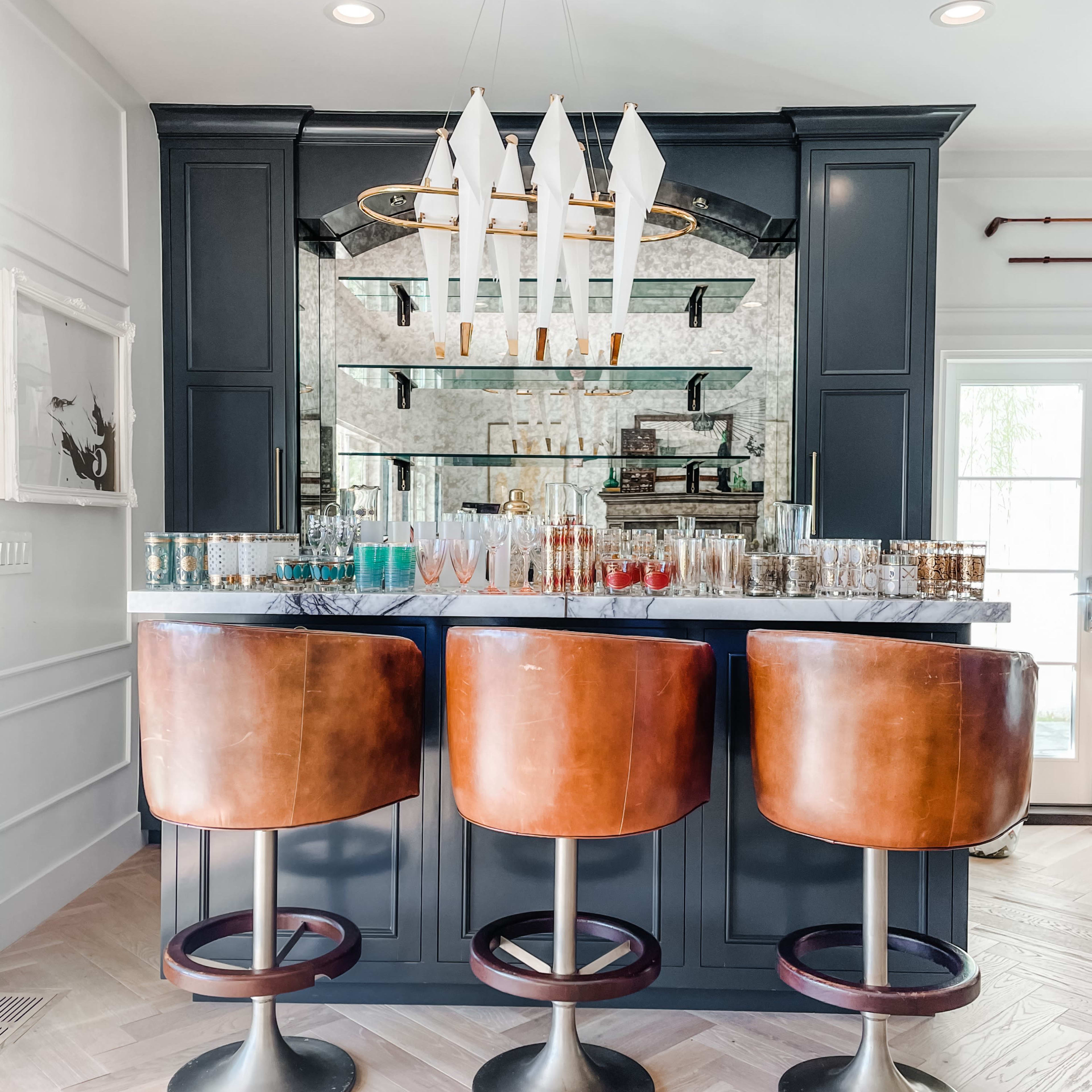 A modern bar area features three brown leather stools in front of a mirrored backsplash and a variety of colorful glassware on the countertop.