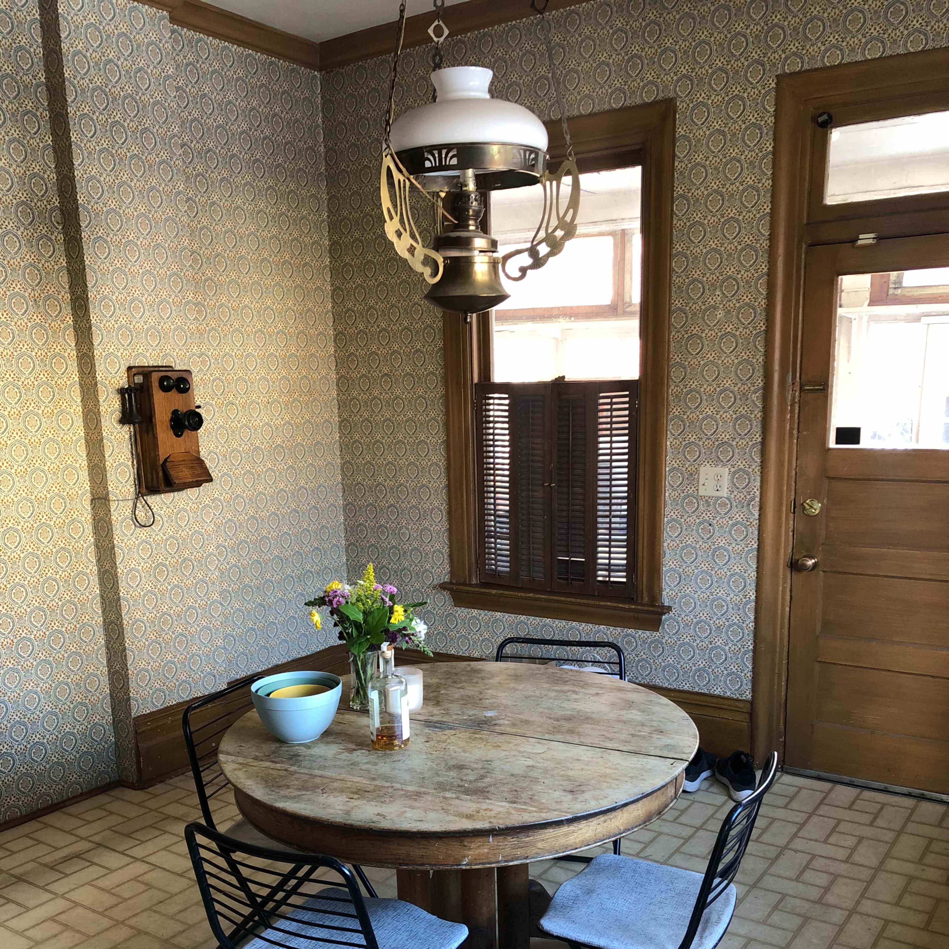 A round wooden dining table with four black metal chairs is set in a vintage kitchen with patterned wallpaper and a hanging lamp.