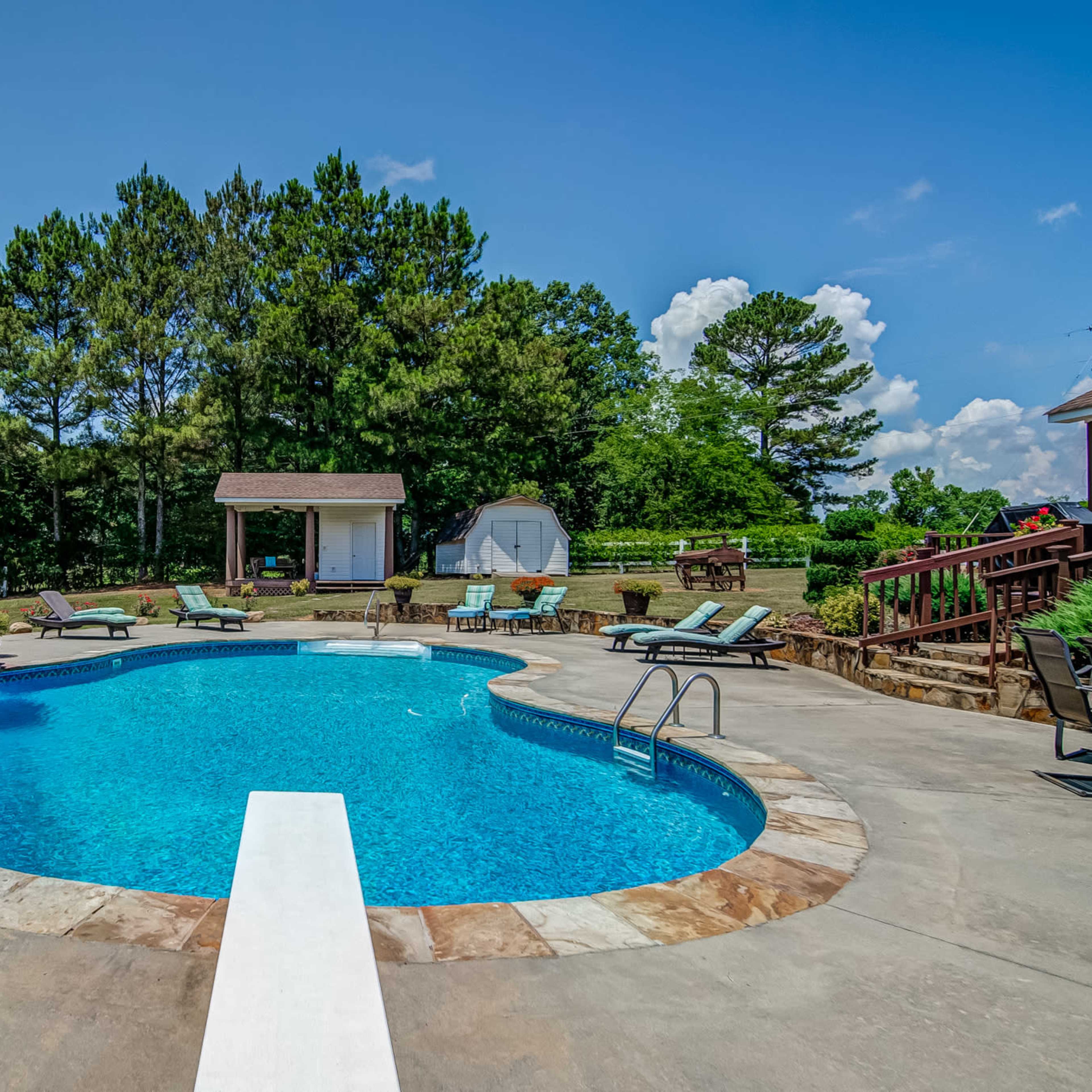 A swimming pool surrounded by lounge chairs and a rock formation in a grassy backyard with trees in the background.