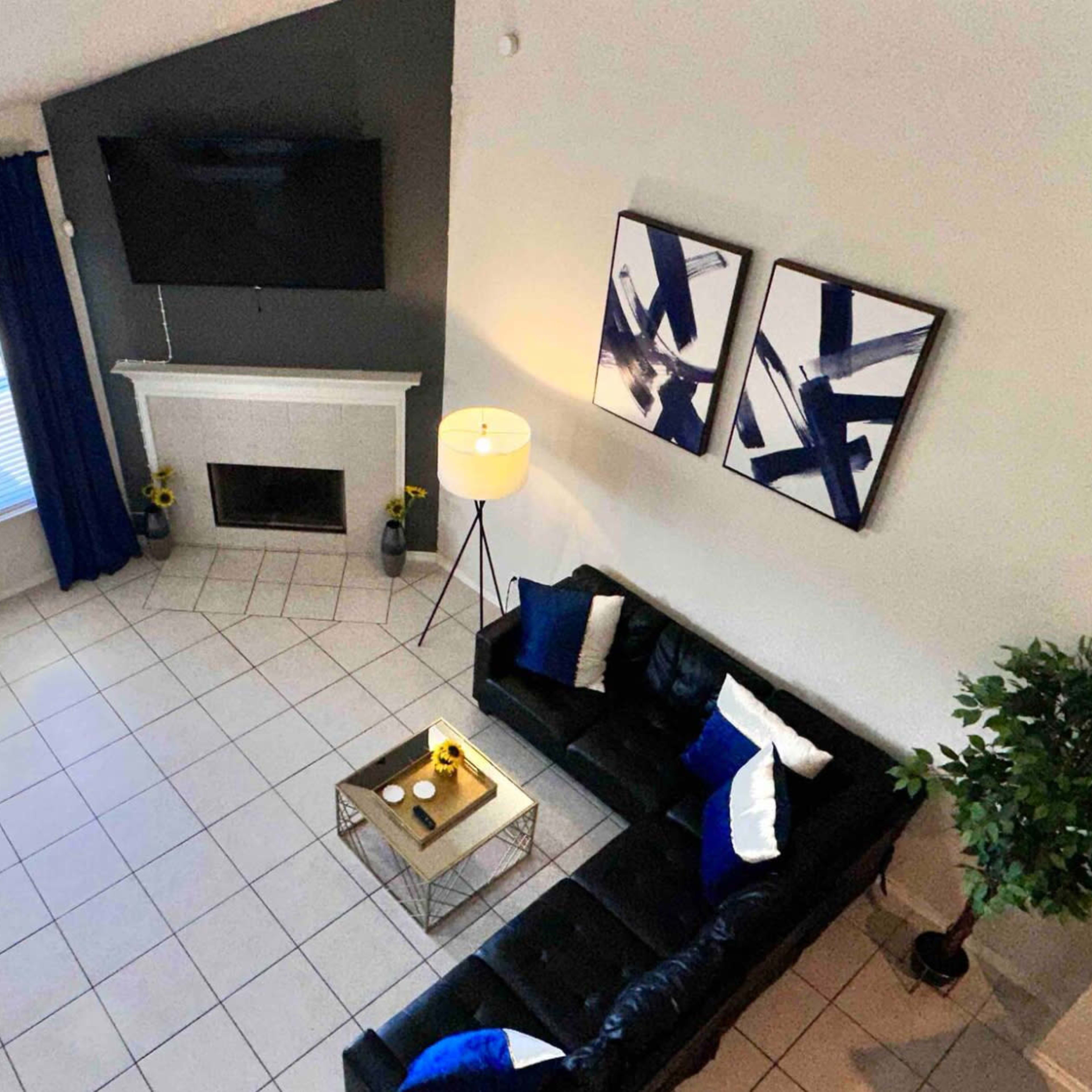 A well-lit living room features a black leather sectional sofa, a glass coffee table, and a fireplace beneath a mounted television, with blue curtains and abstract wall art.
