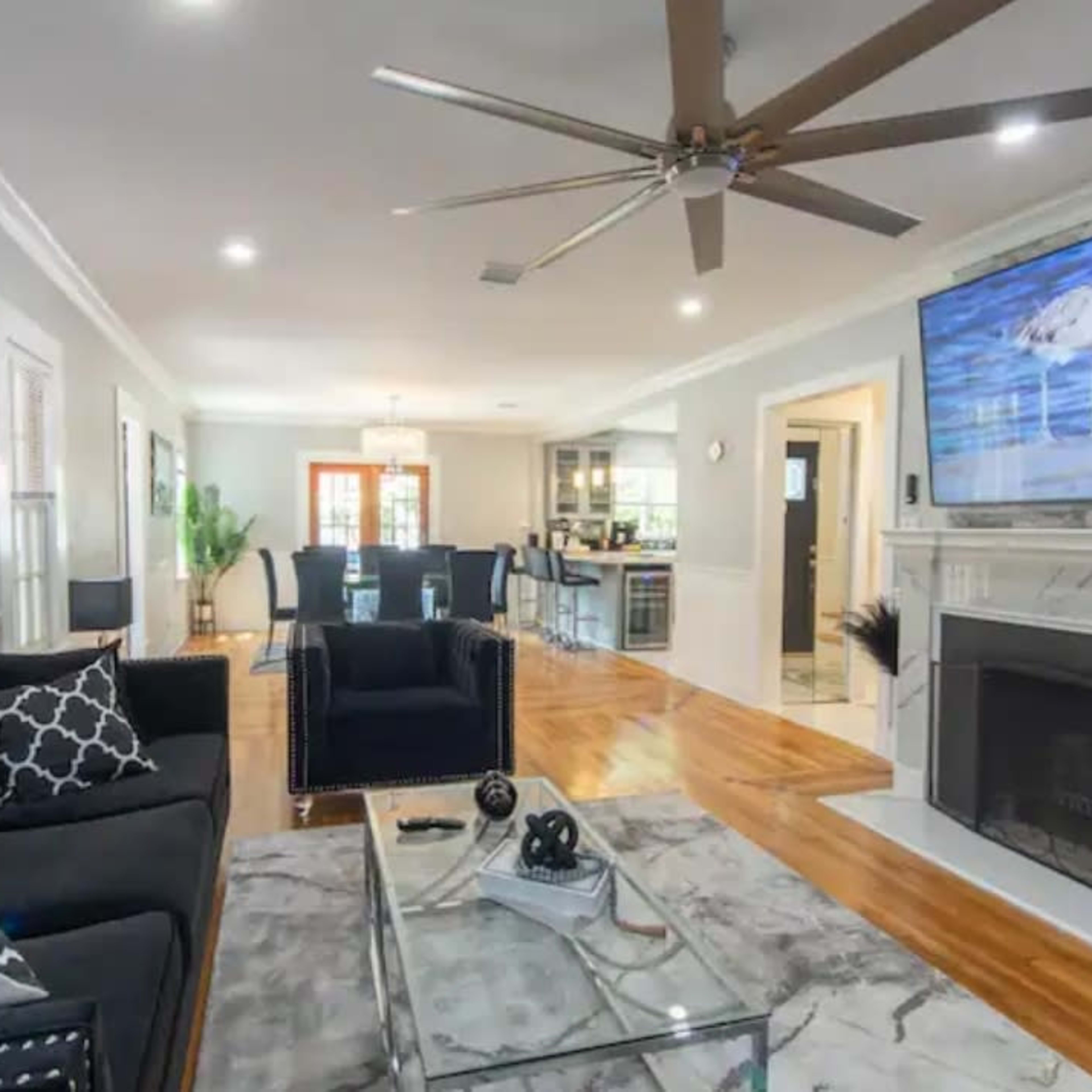 The living room features a large black sofa, a glass coffee table, and a mounted TV above a fireplace, with a view into a dining area and modern kitchen.