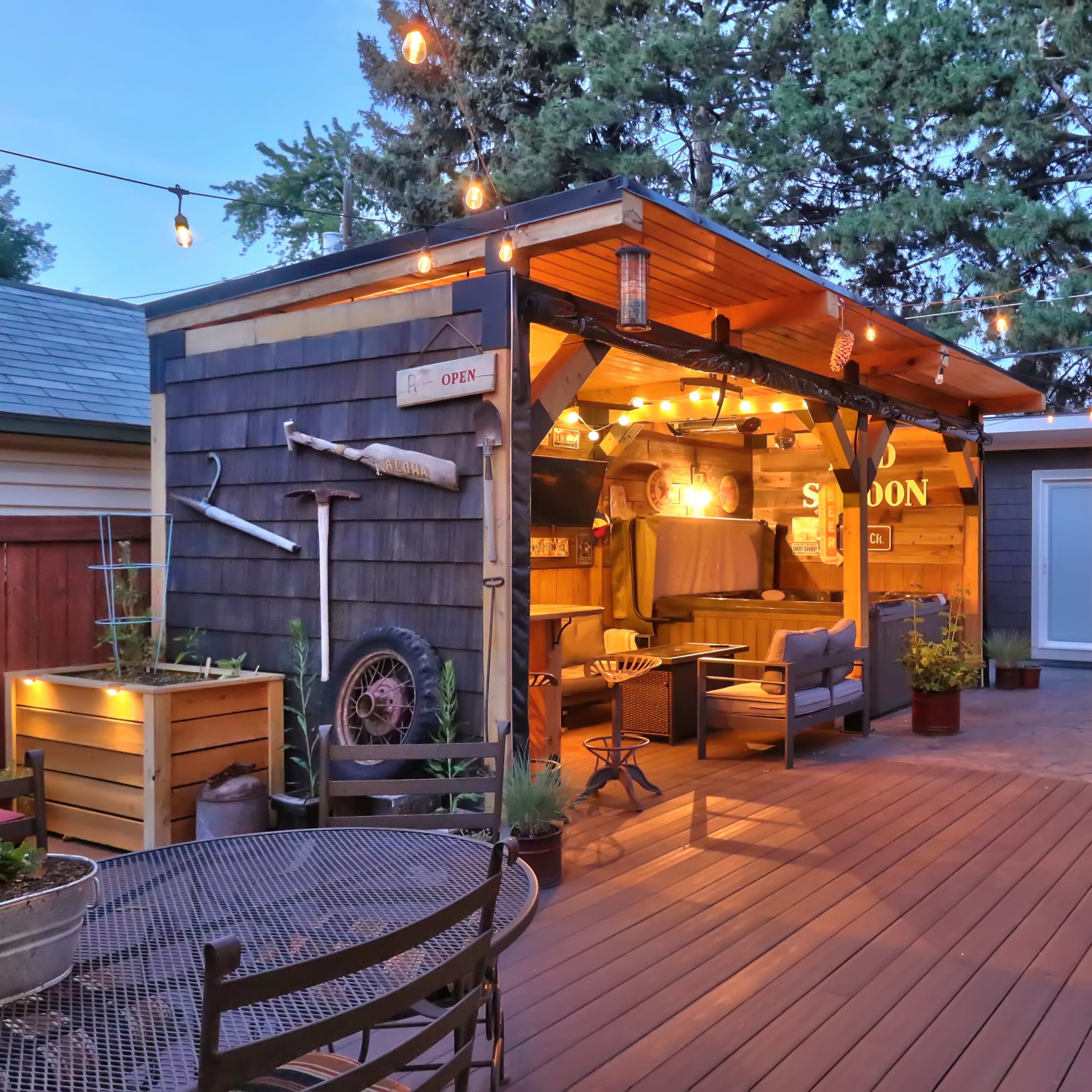 A wooden deck features a well-lit outdoor lounge area with a covered seating space and decorative string lights, alongside a small dining table.