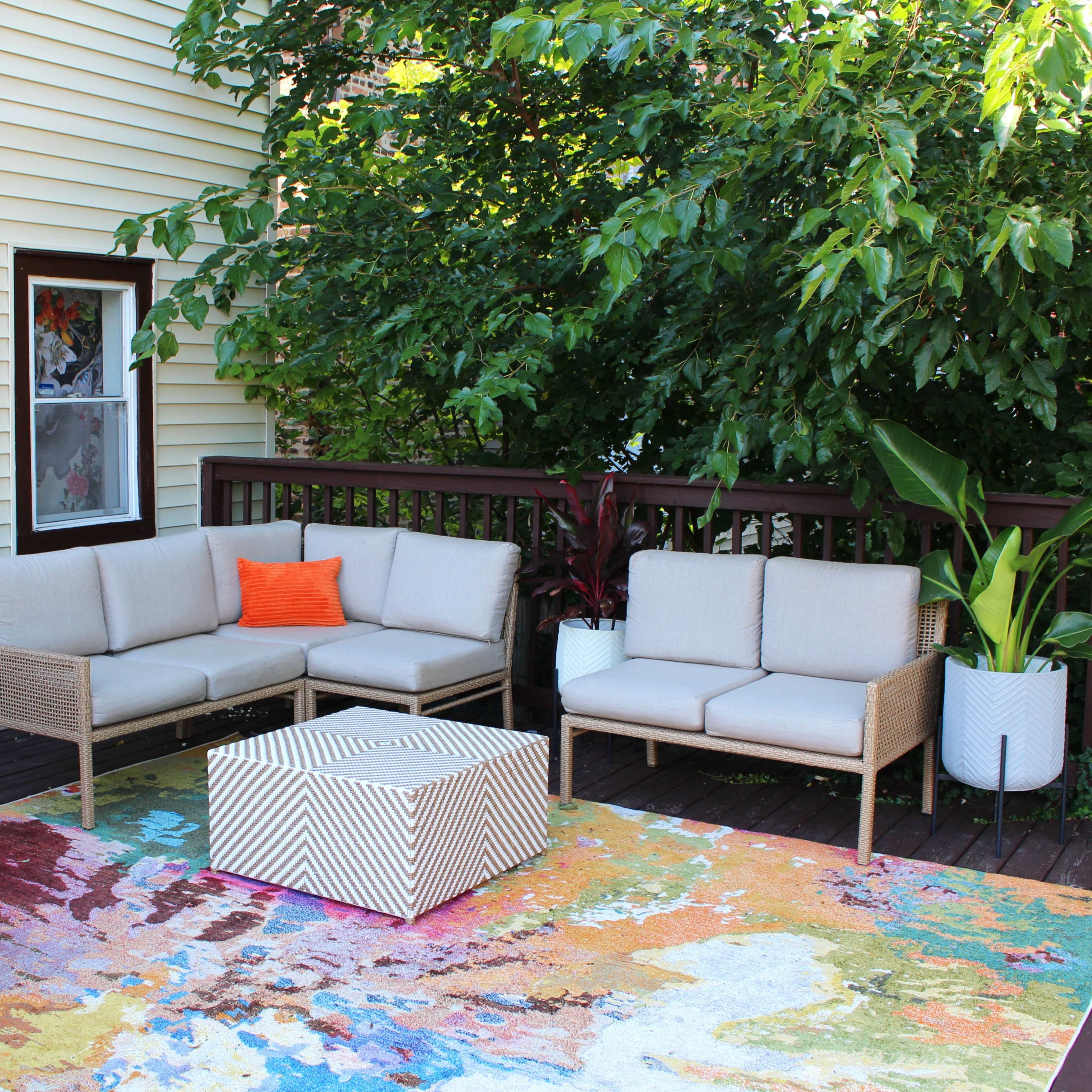 A cozy outdoor seating area features a light-colored sofa set with an orange accent pillow, a striped coffee table, and potted plants on a colorful area rug.