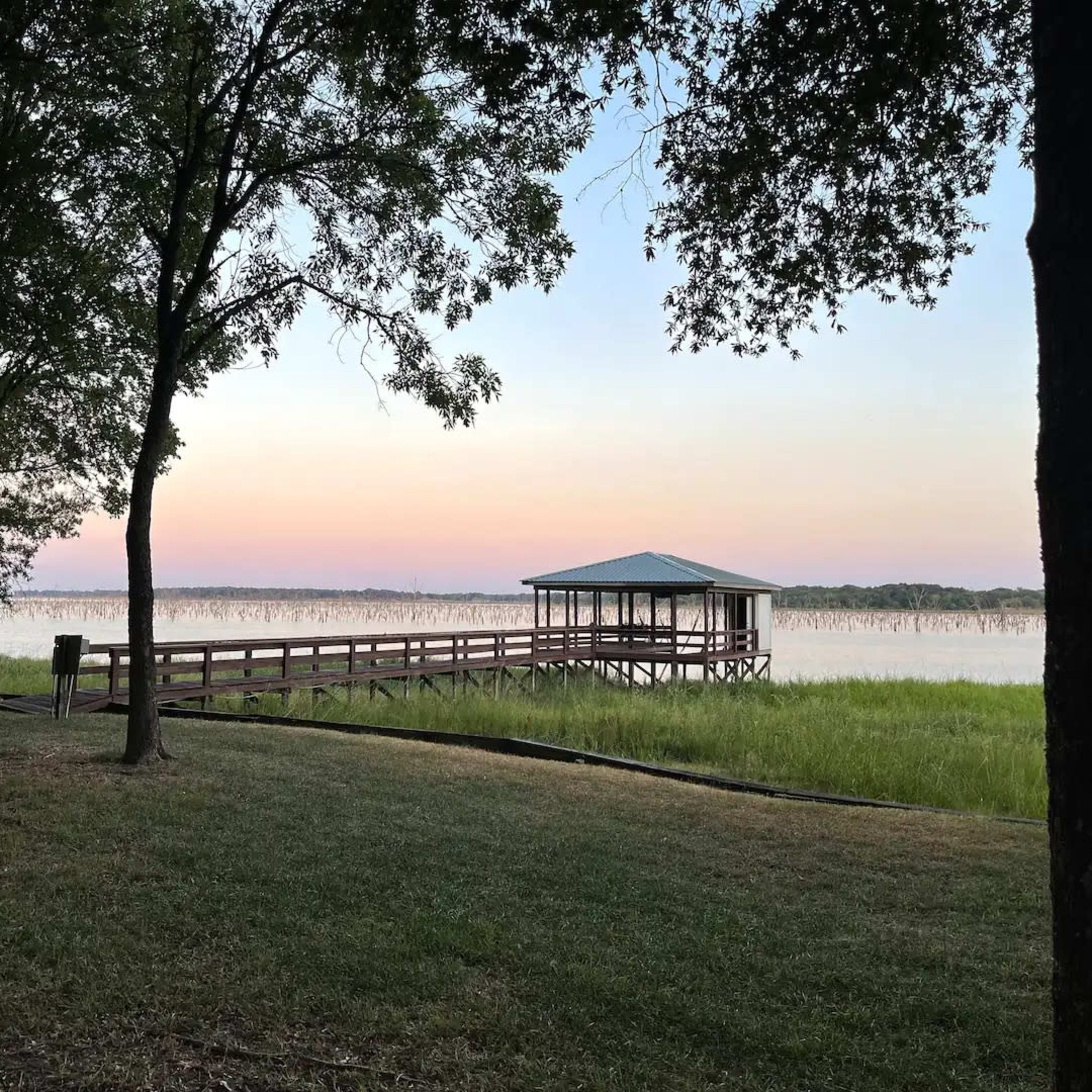 A wooden dock extends over a calm body of water surrounded by trees at dusk.