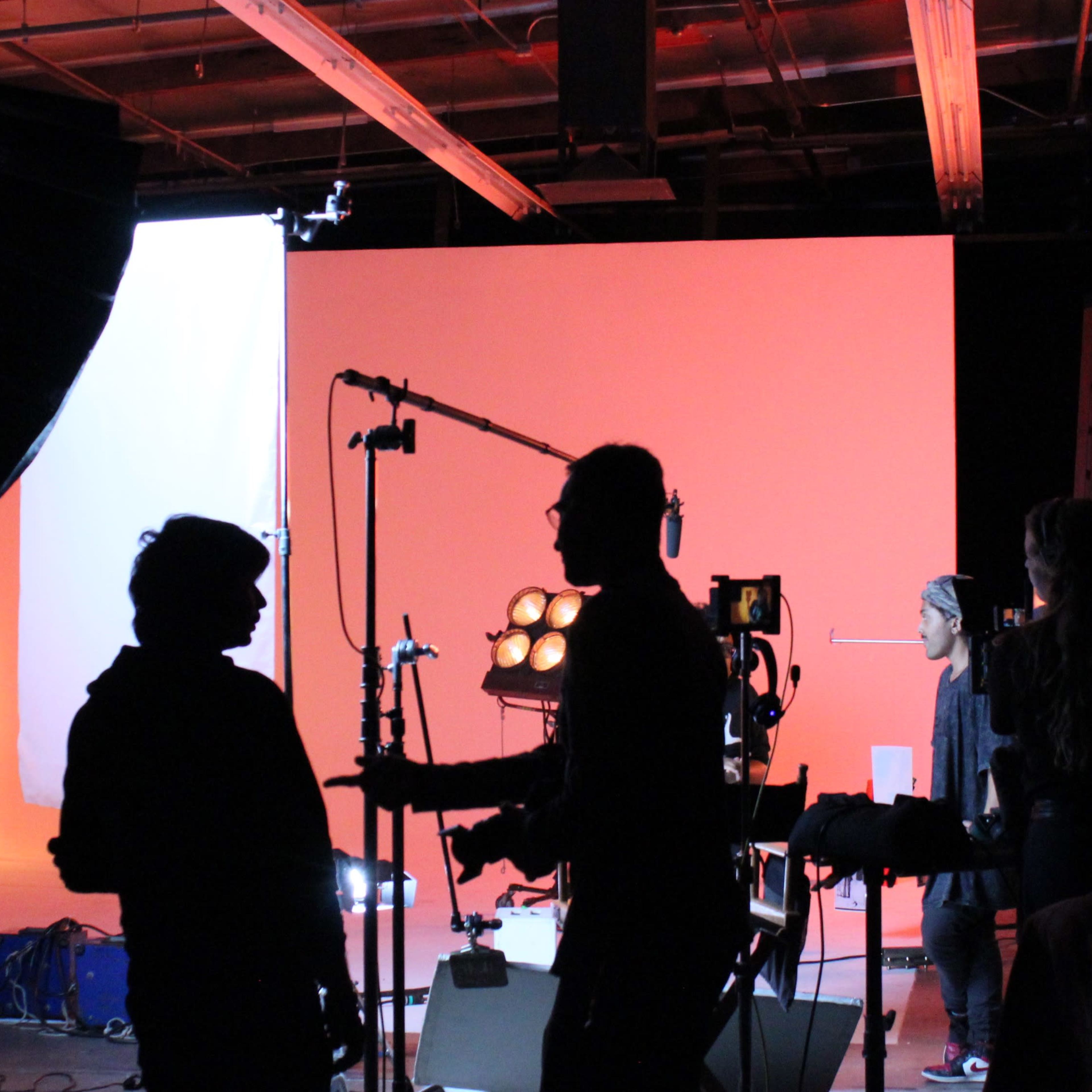 A film set is illuminated by various lights, with silhouettes of crew members working in front of a brightly colored backdrop.